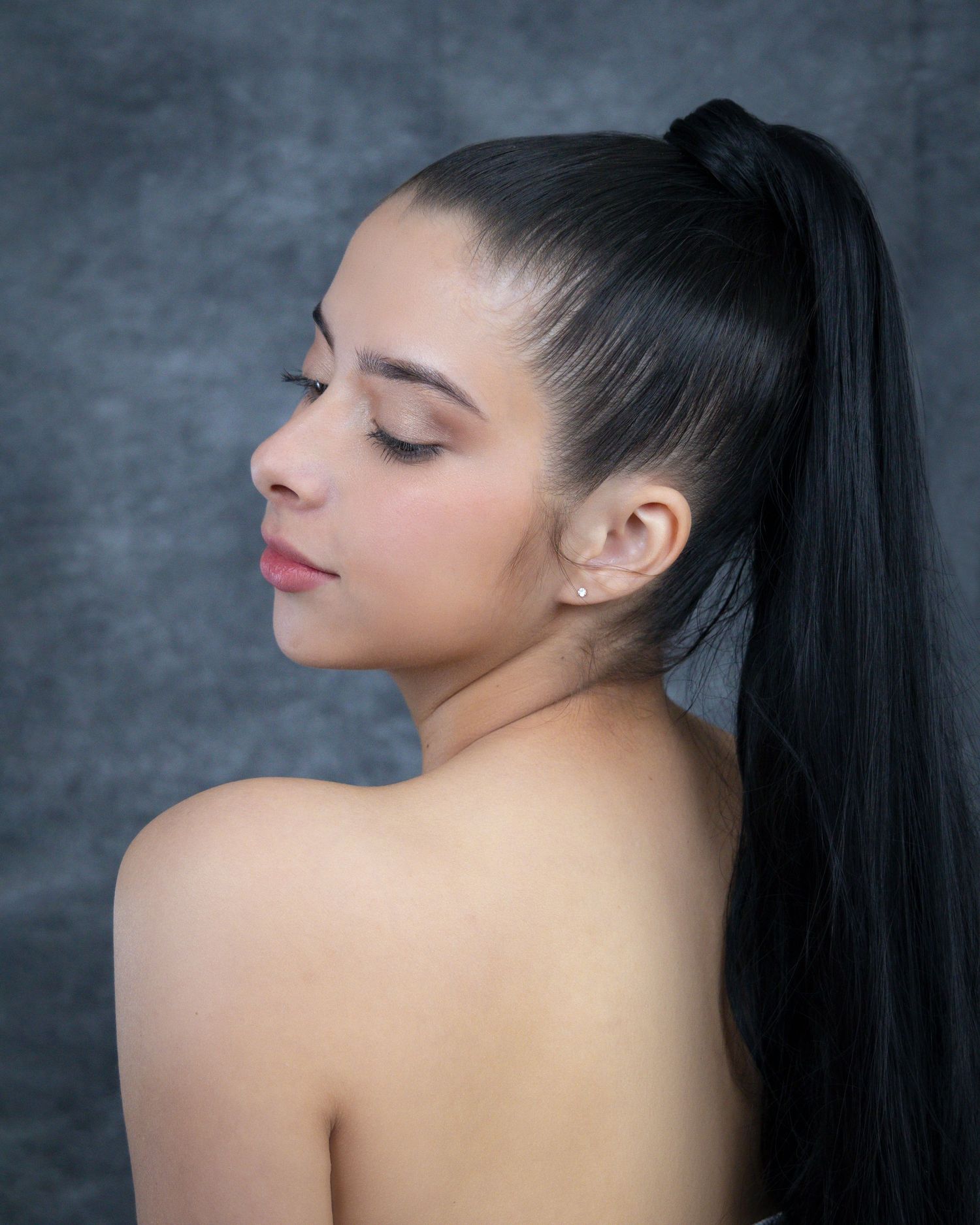 Young woman with long black ponytail poses in profile view, eyes closed, wearing small stud earrings against gray backdrop.