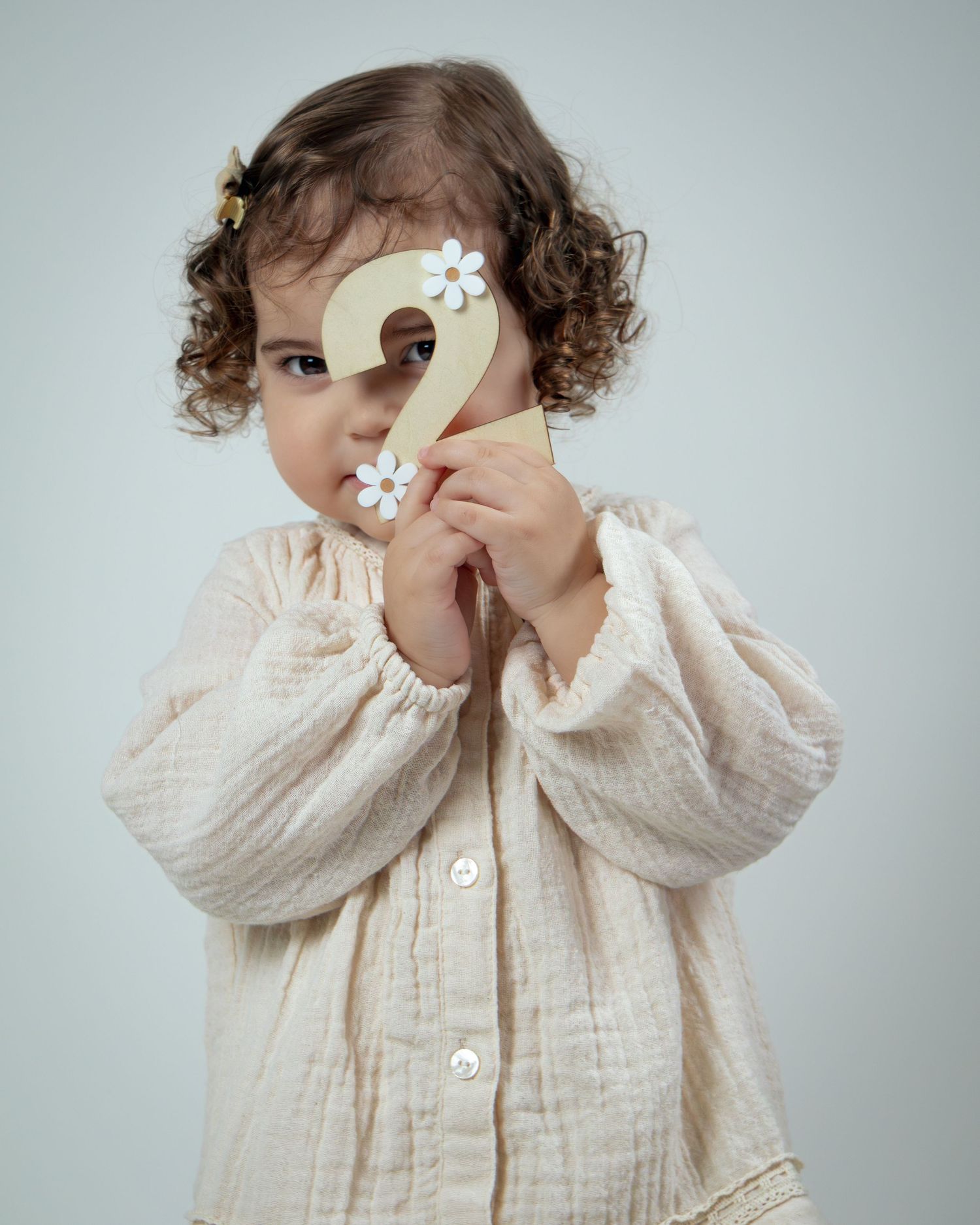 A toddler in a cream cotton shirt holding up a wooden number 2 sign decorated with white flowers.