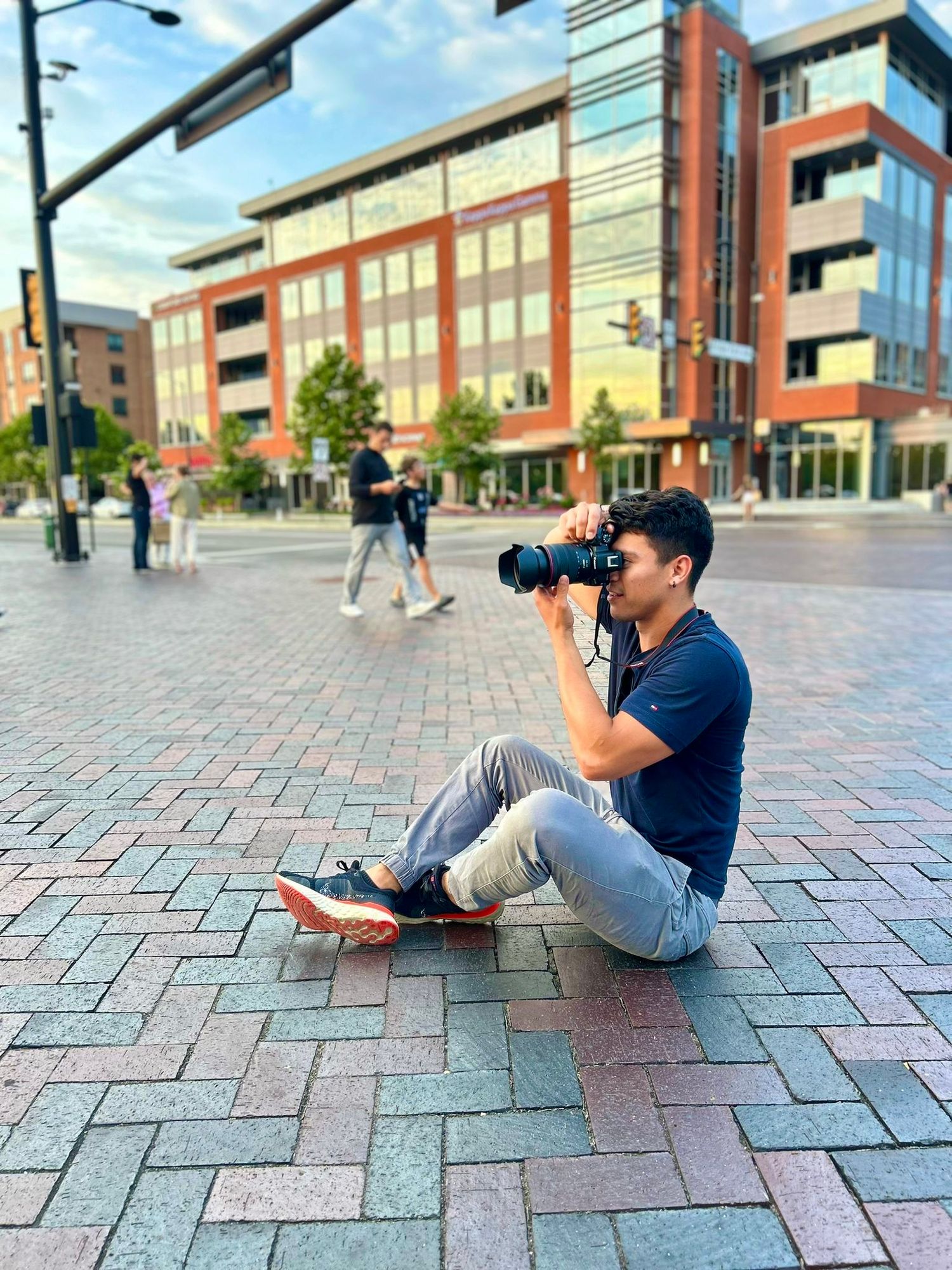 Photographer sitting on brick pavement taking pictures in front of modern building complex.