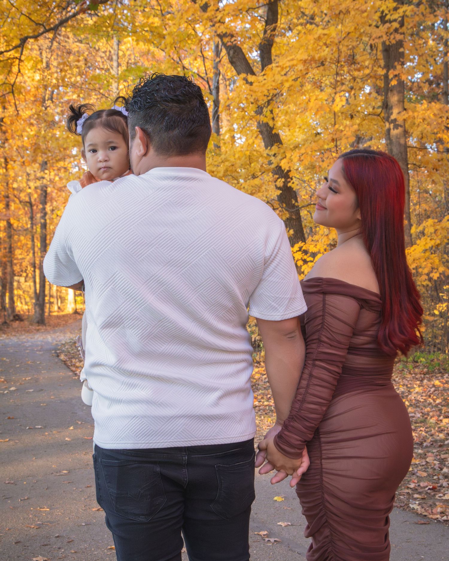 Family of three poses on autumn path, man holding toddler beside red-haired woman in brown dress.