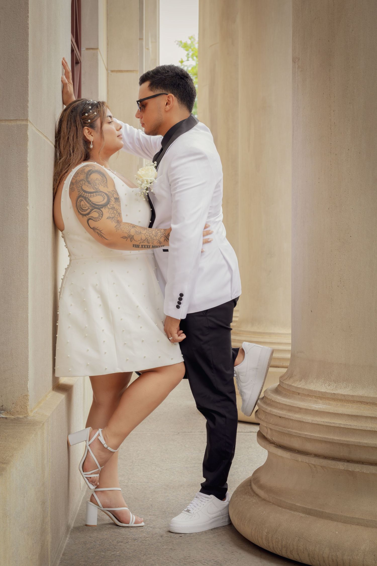 Couple in white wedding attire sharing a romantic moment between stone columns, bride with tattoos in mini dress and groom in white blazer.