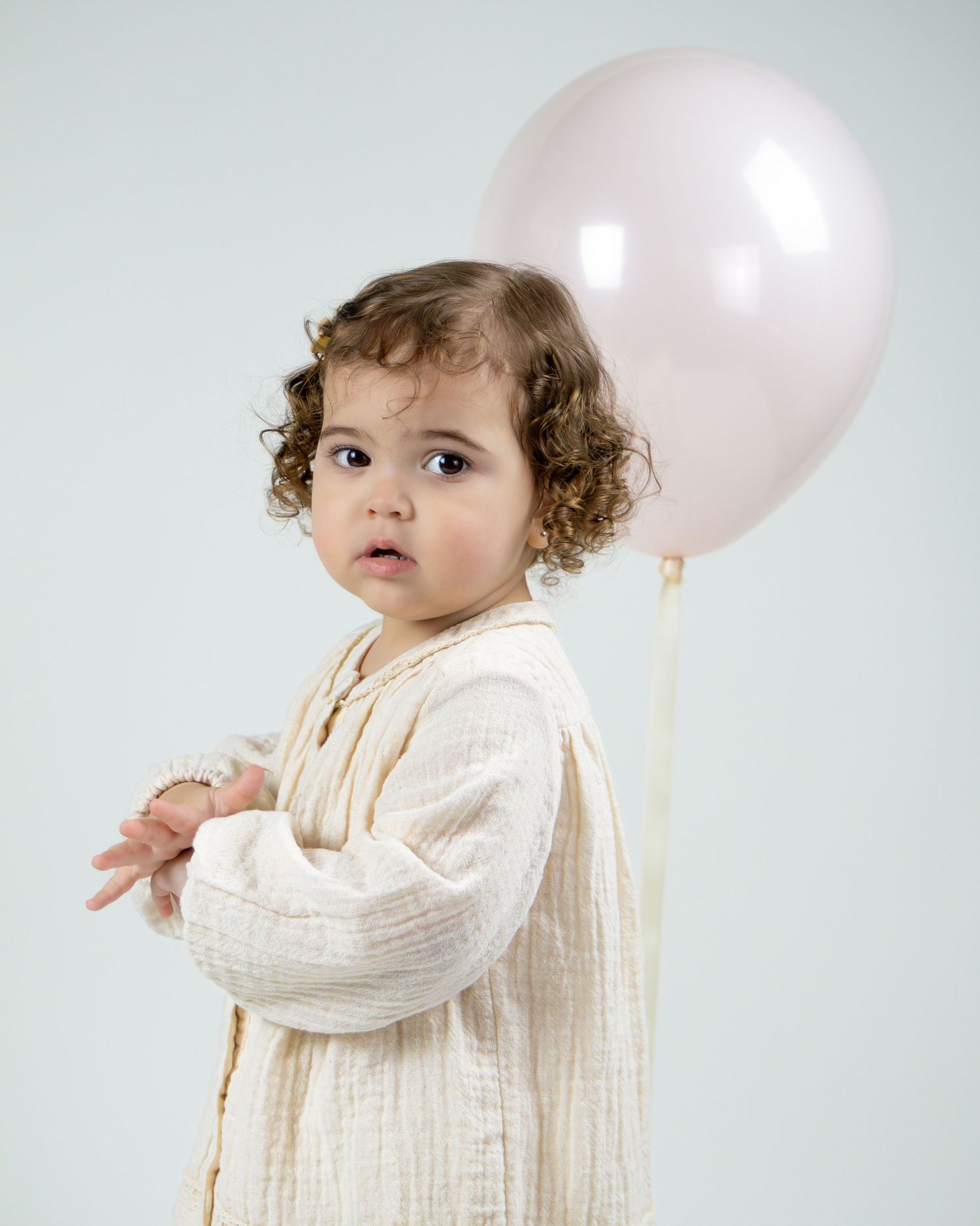 A small figure in a flowing cream dress poses with a soft pink balloon against a light background.