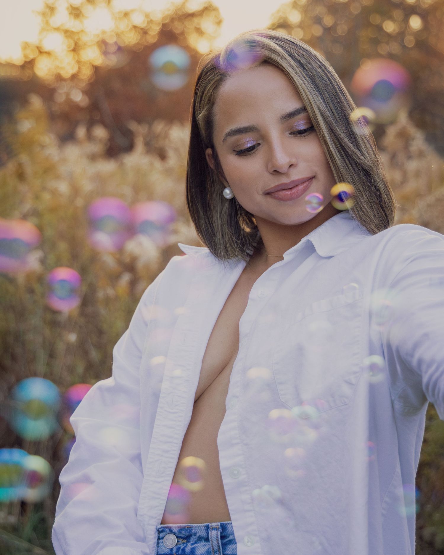 Young woman in open white shirt smiling with eyes closed, surrounded by colorful soap bubbles in golden sunlight.