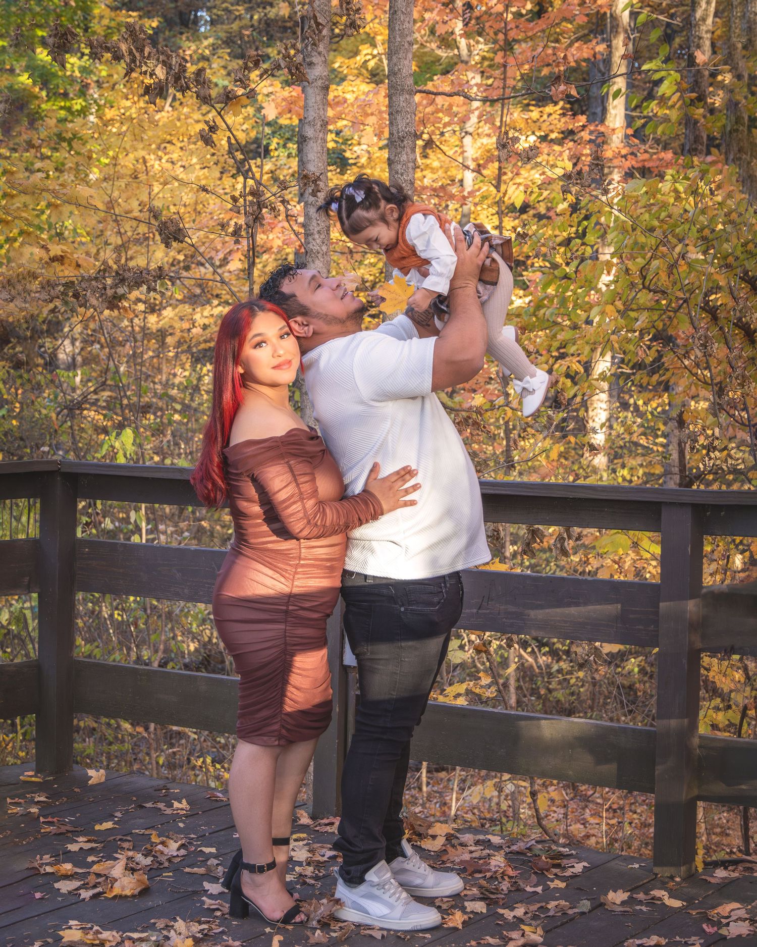 A couple poses on a wooden bridge in autumn, man lifting a baby girl while woman in brown dress leans against him.