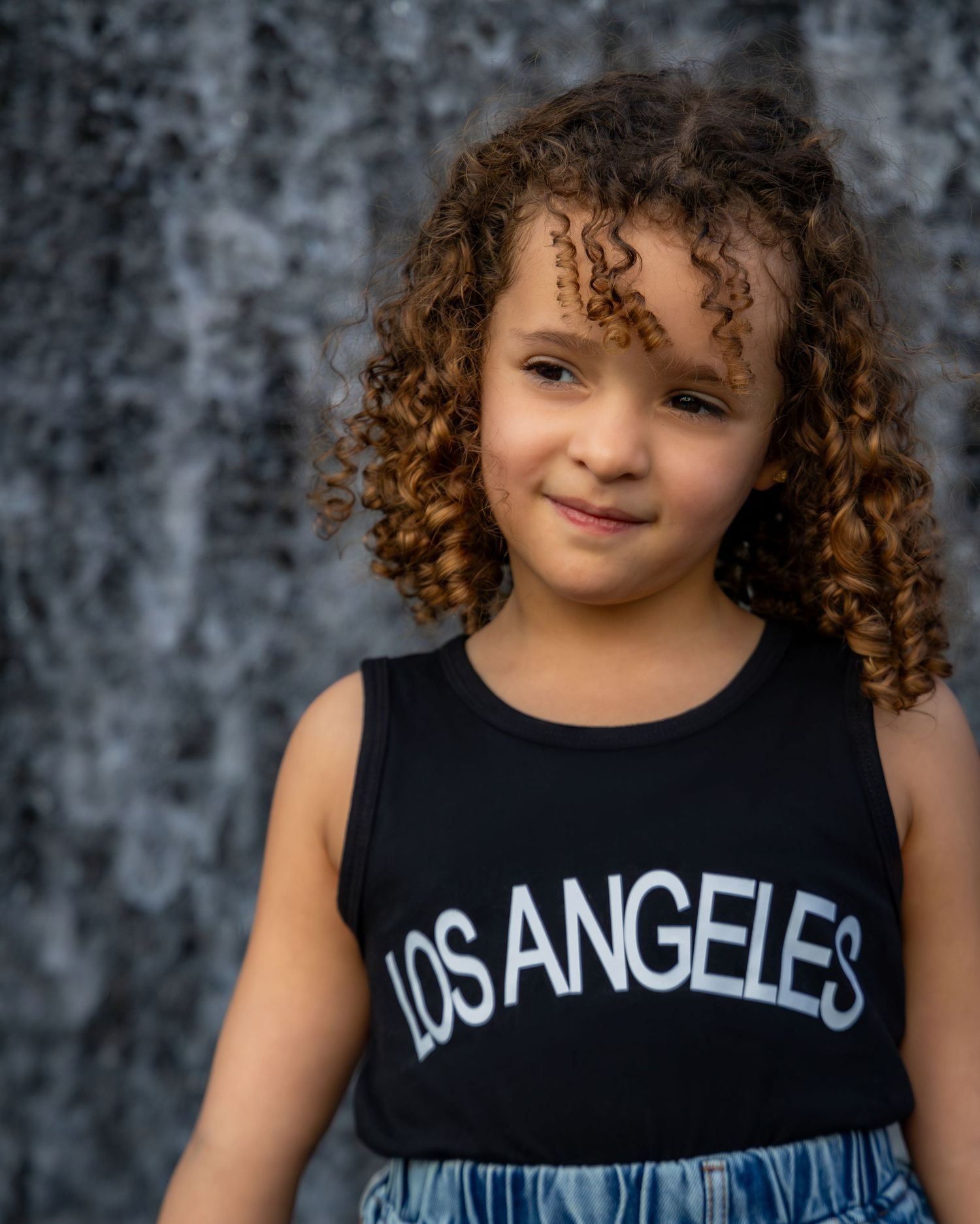 Young girl with curly hair wearing a black Los Angeles tank top, smiling in front of a waterfall backdrop.