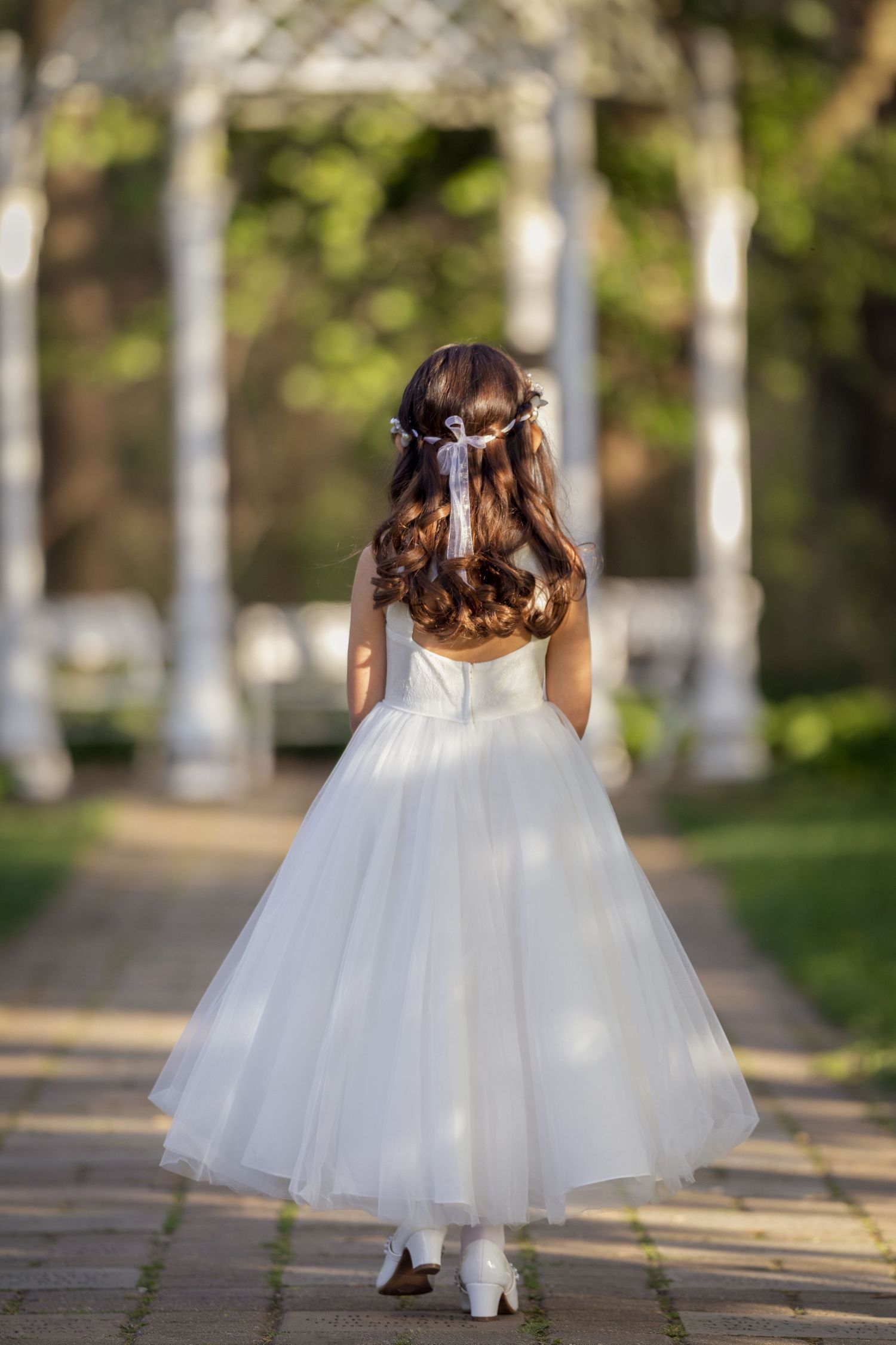 Young girl in white tulle dress with floral hair accessories walks garden path, viewed from behind.