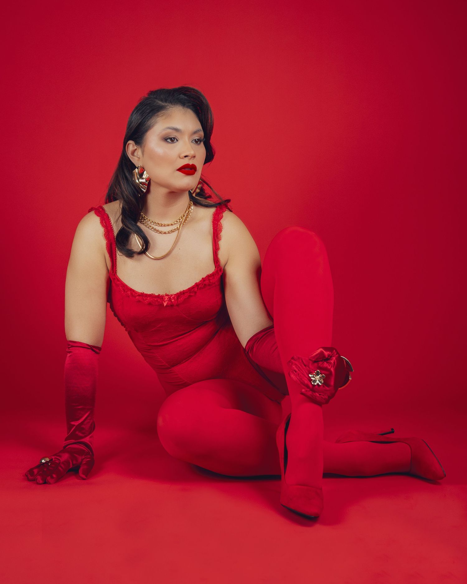 Woman in red jumpsuit, gloves, and stockings poses on floor against red background, wearing gold jewelry and red lipstick.