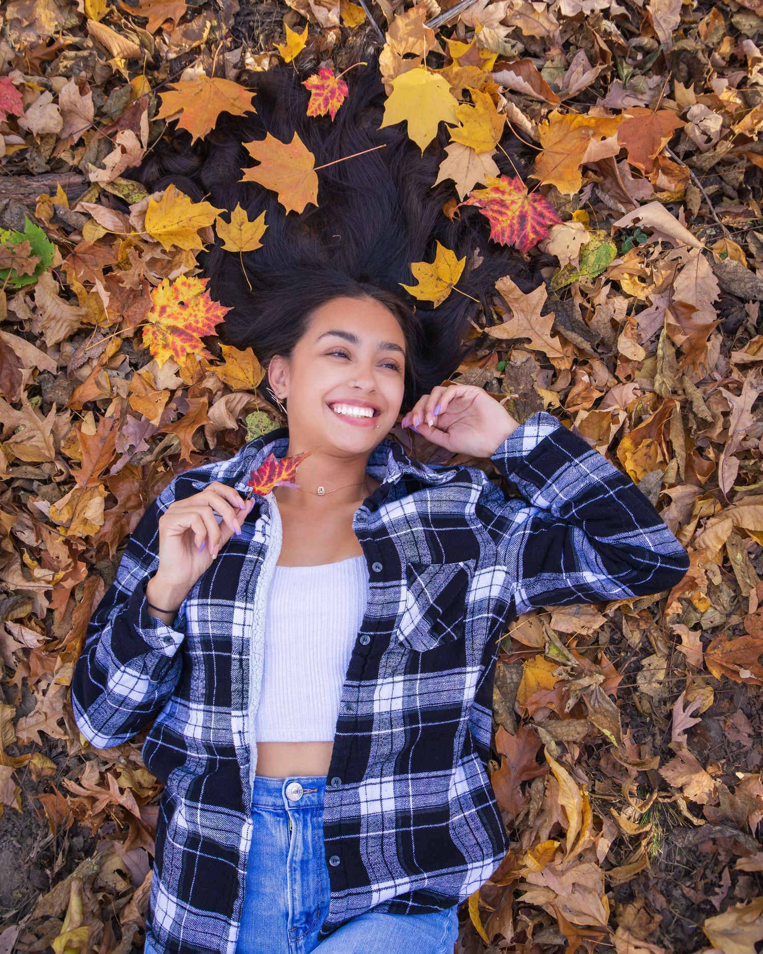 Young woman smiling while lying on colorful autumn leaves, wearing a plaid shirt and jeans in a fall outdoor setting.