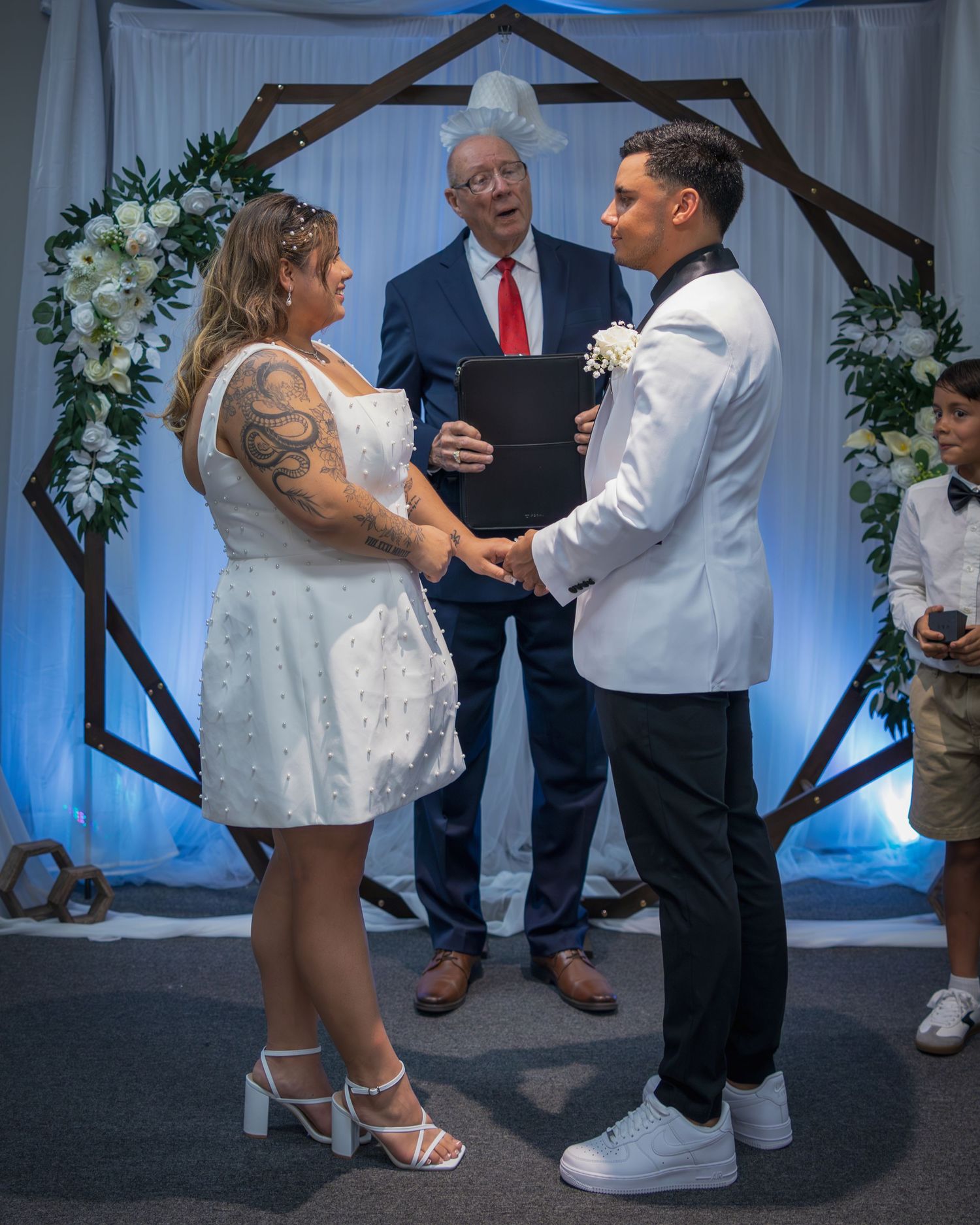 Couple exchanging vows at wedding ceremony before officiant under geometric wooden arch with white floral decorations.