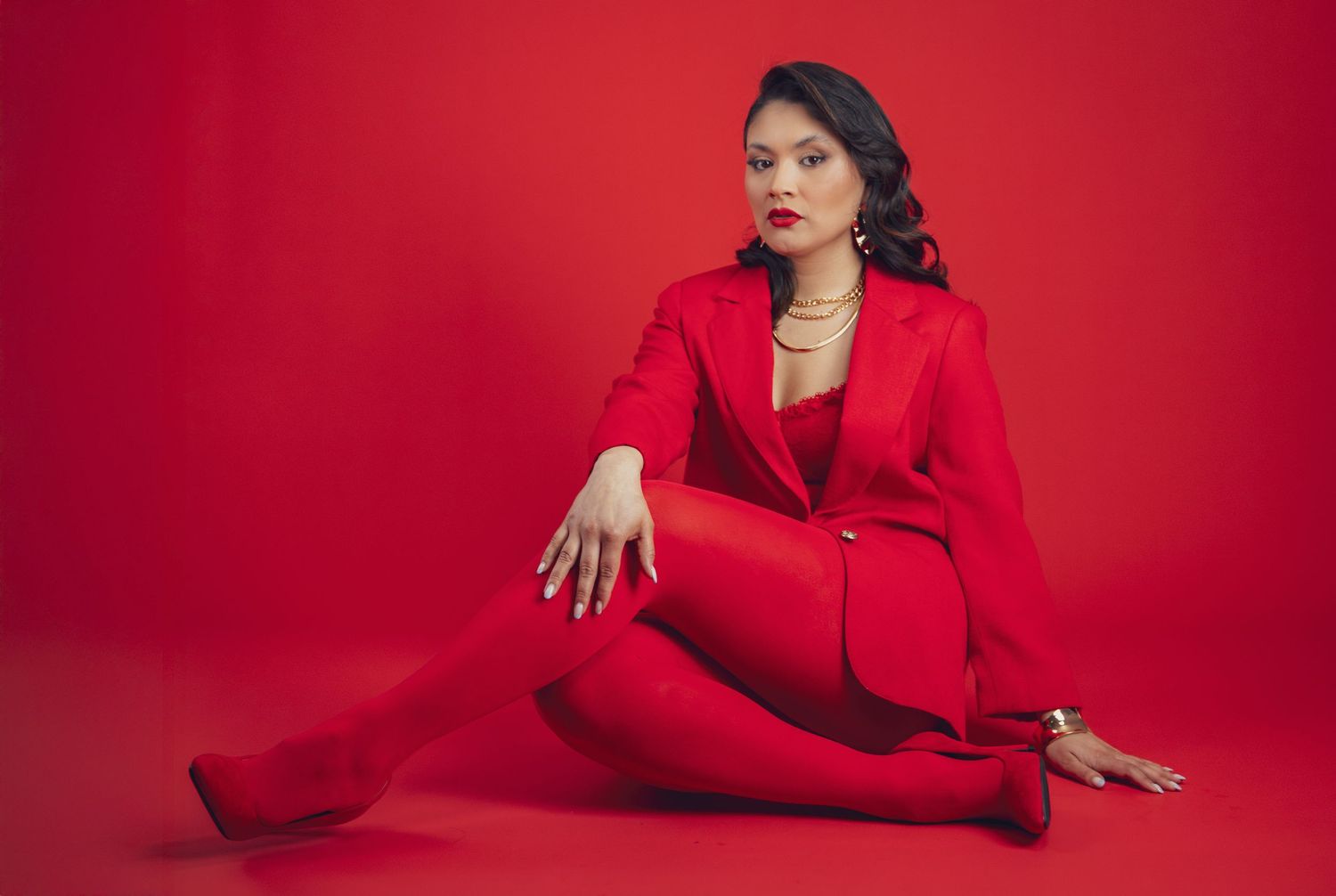 Woman in red suit sitting on floor against red background, wearing heels and gold jewelry.