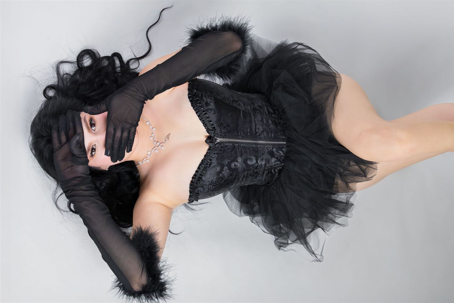 Woman in black corset, tutu skirt, and long gloves posing dramatically on white background.