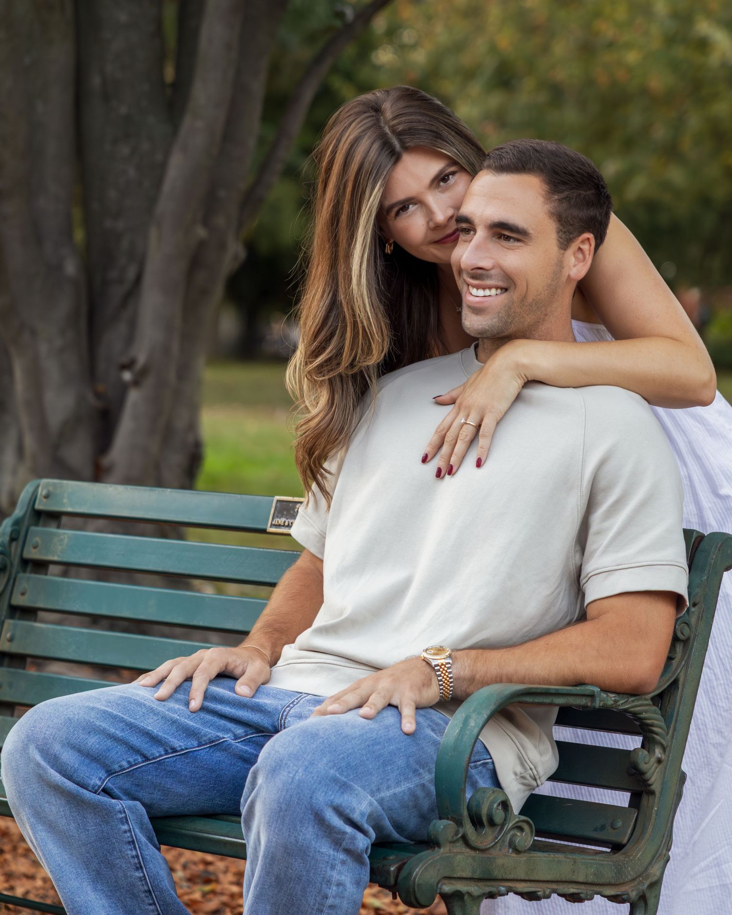 A smiling couple poses on a green park bench, woman hugging man from behind, both wearing casual outfits outdoors.
