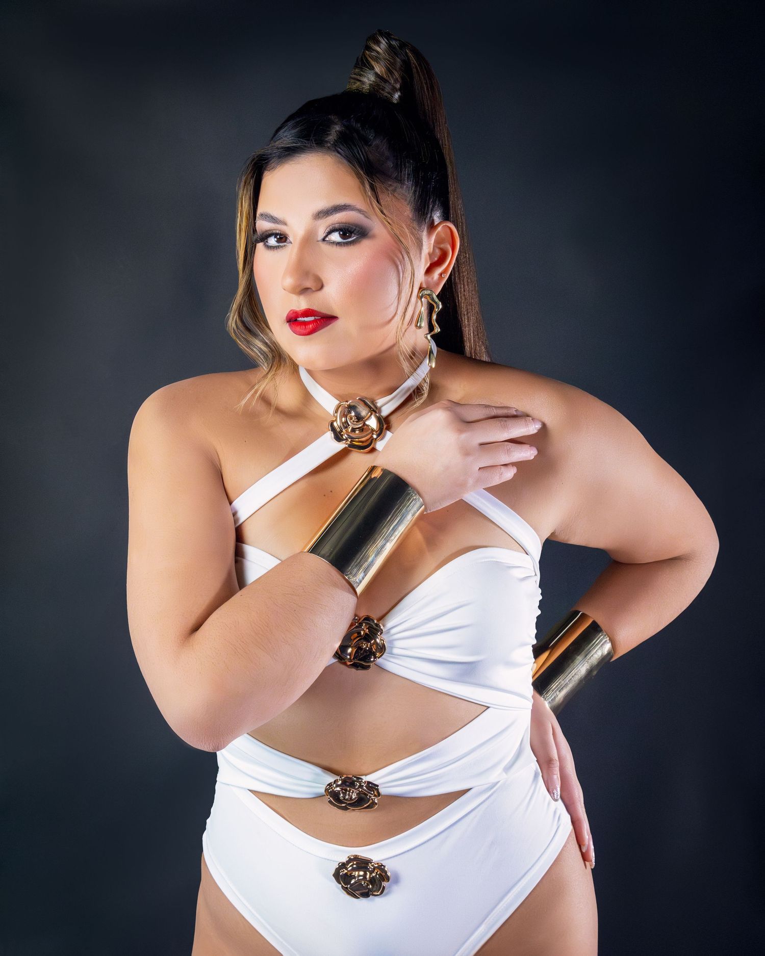 Woman in white cutout bodysuit with gold cuff bracelets and choker necklace poses against dark background.