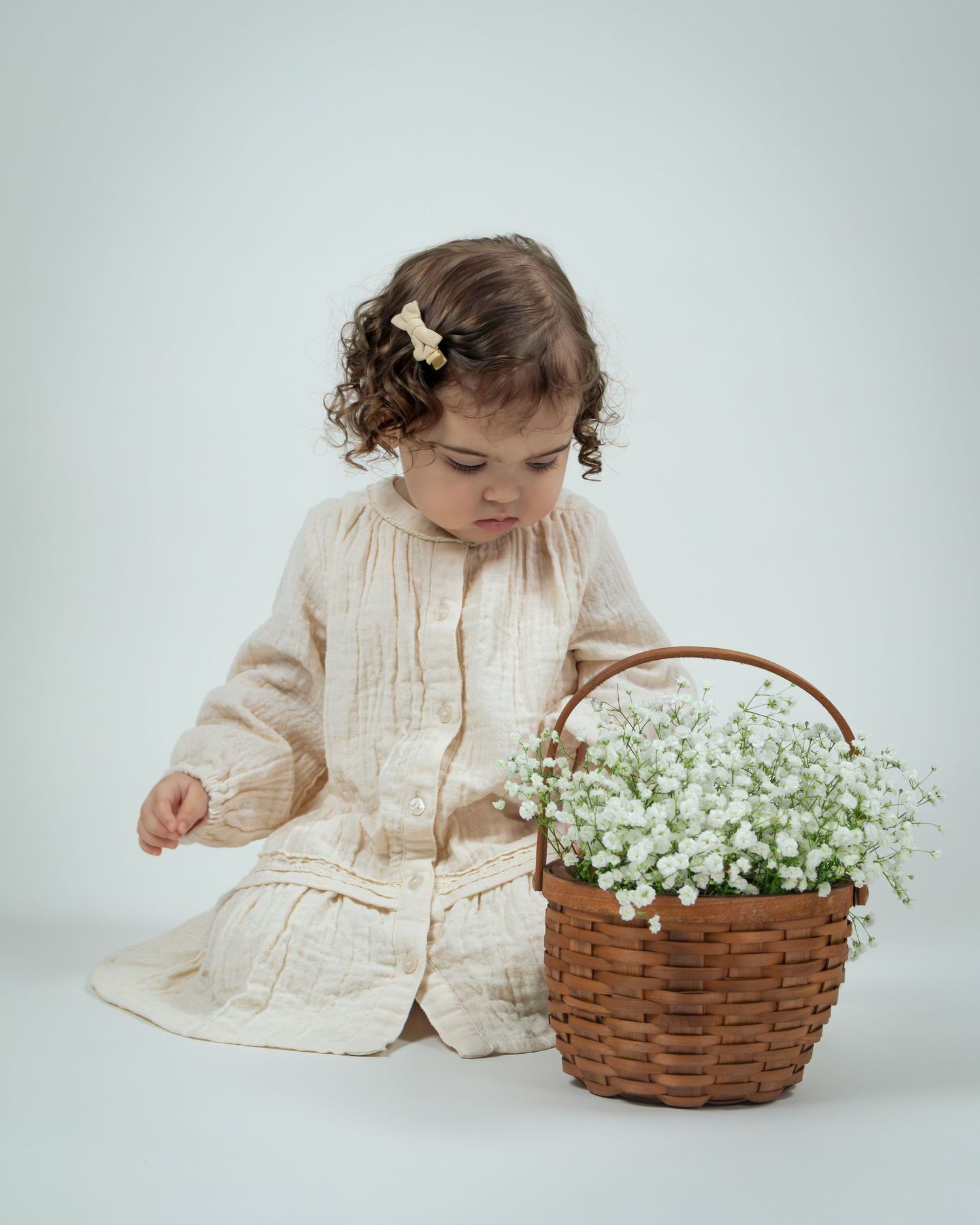 Toddler girl in cream dress sitting beside a wicker basket of white flowers, with a floral hair clip.