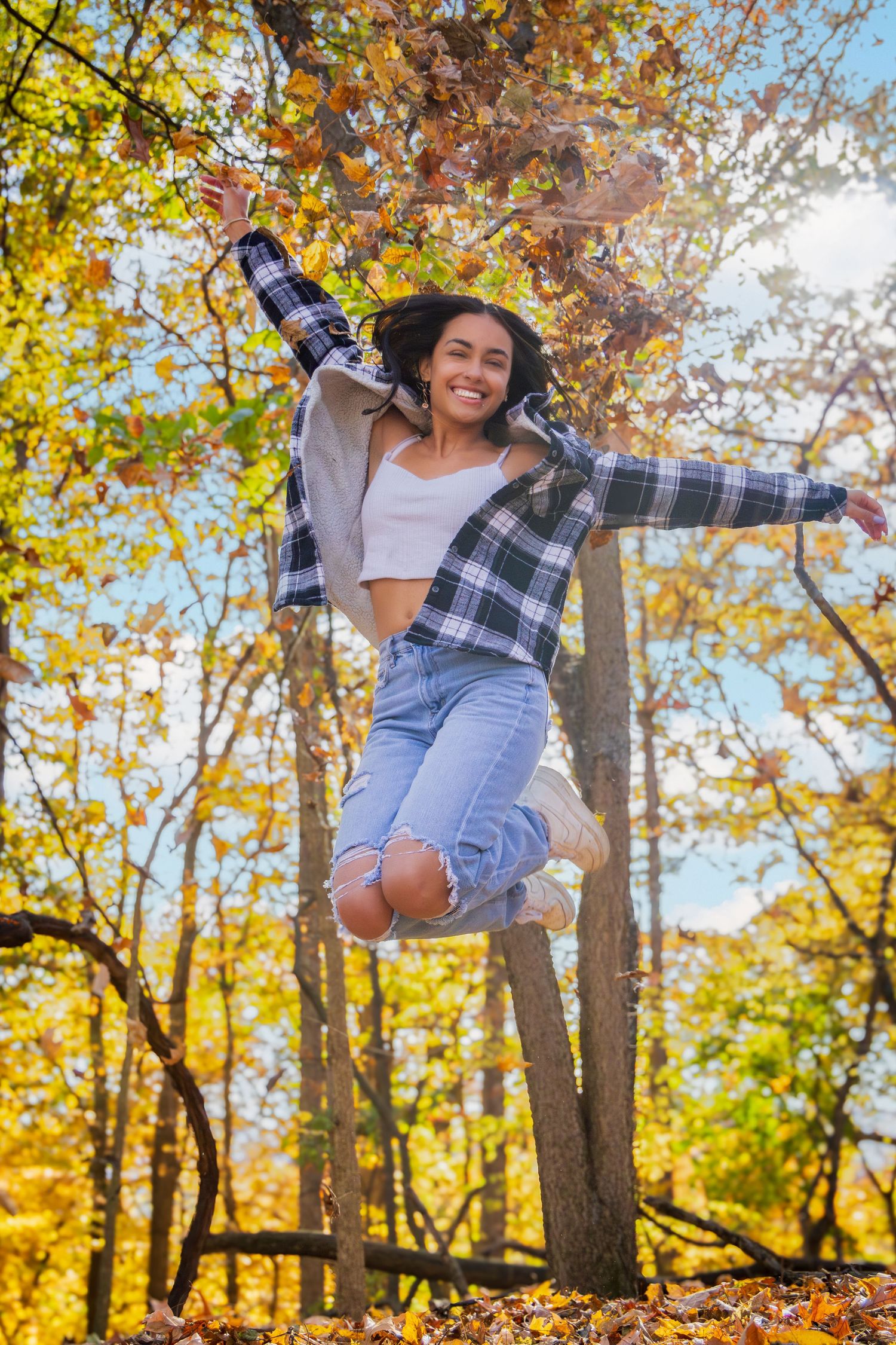 Person in plaid shirt and jeans jumping joyfully among autumn trees with fallen leaves.