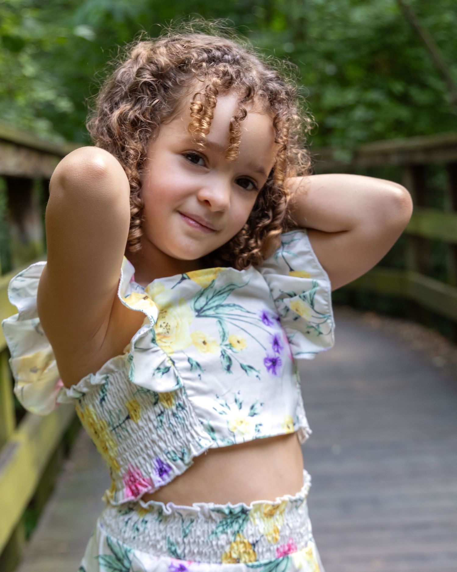 Child in floral two-piece outfit poses playfully with hands behind head outdoors.