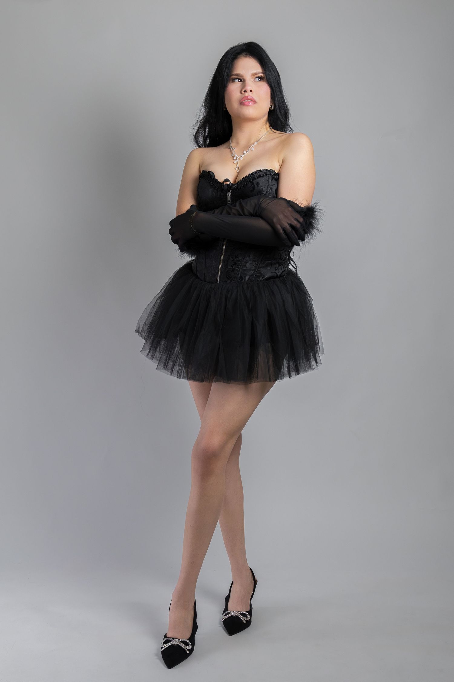 Woman in black strapless corset mini tulle skirt, long gloves, and heels posing against gray background.