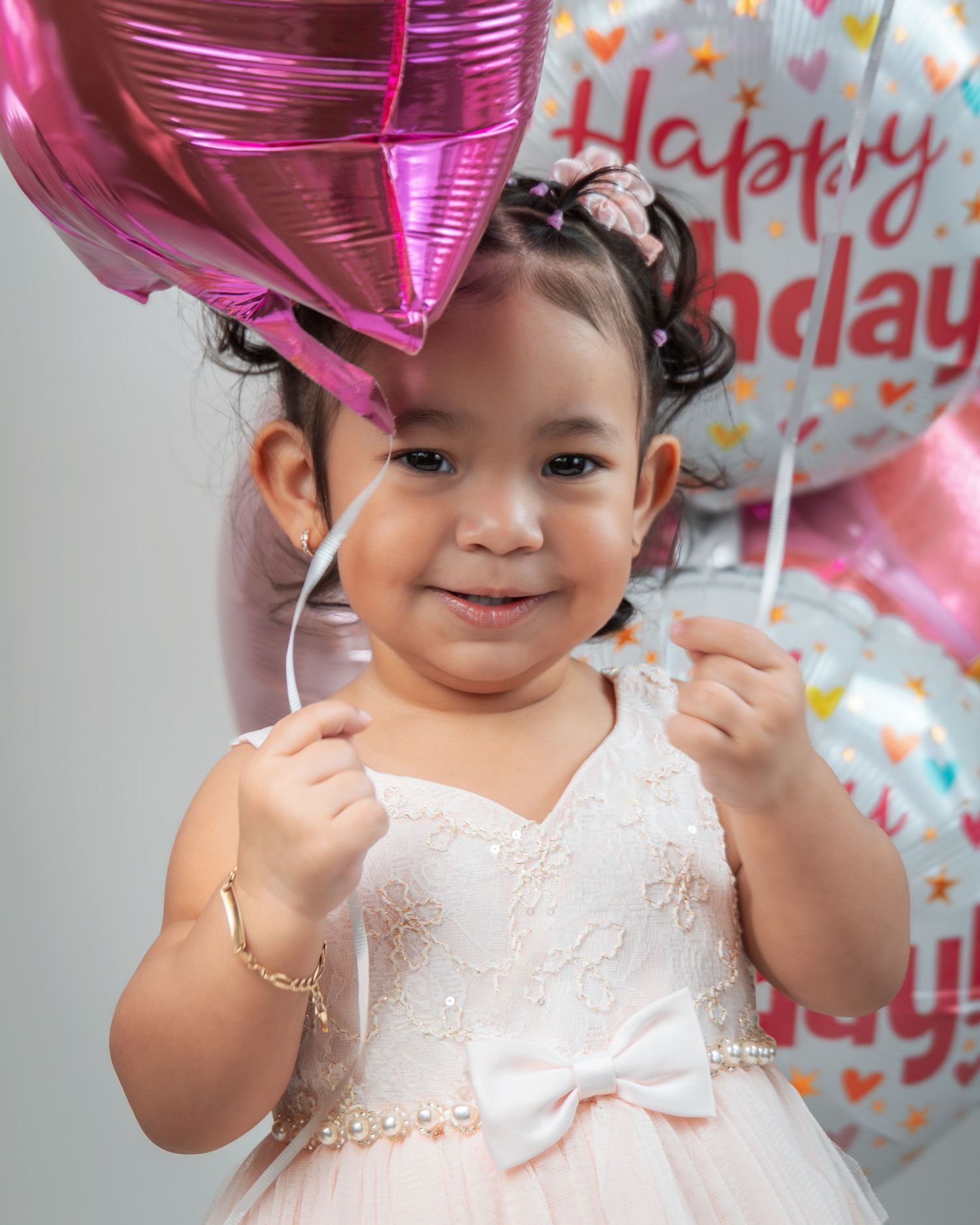 A small child in a white dress holds pink birthday balloons during a celebration with decorative party backgrounds.
