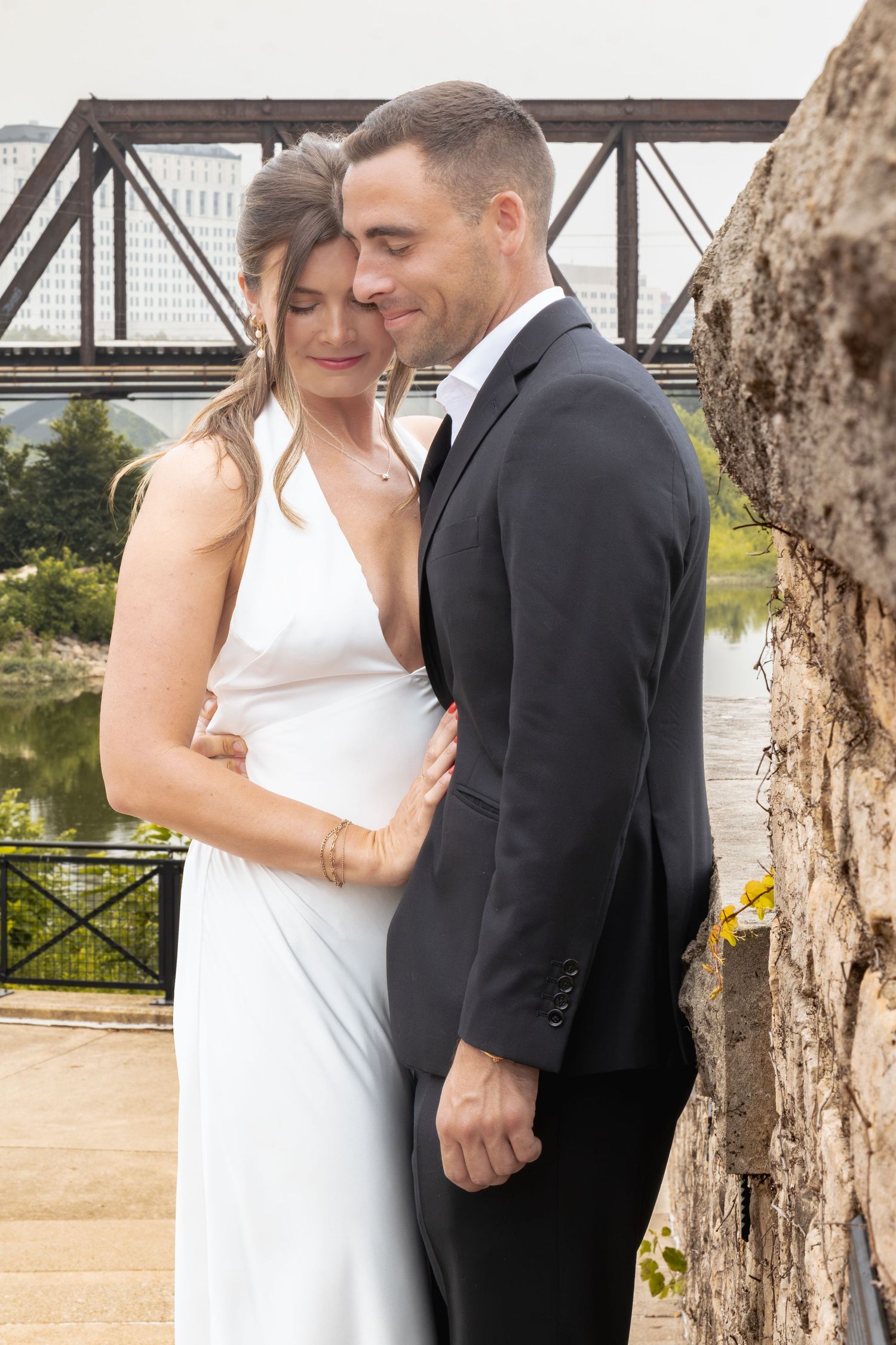 Couple in wedding attire embracing near stone wall with bridge and river backdrop.