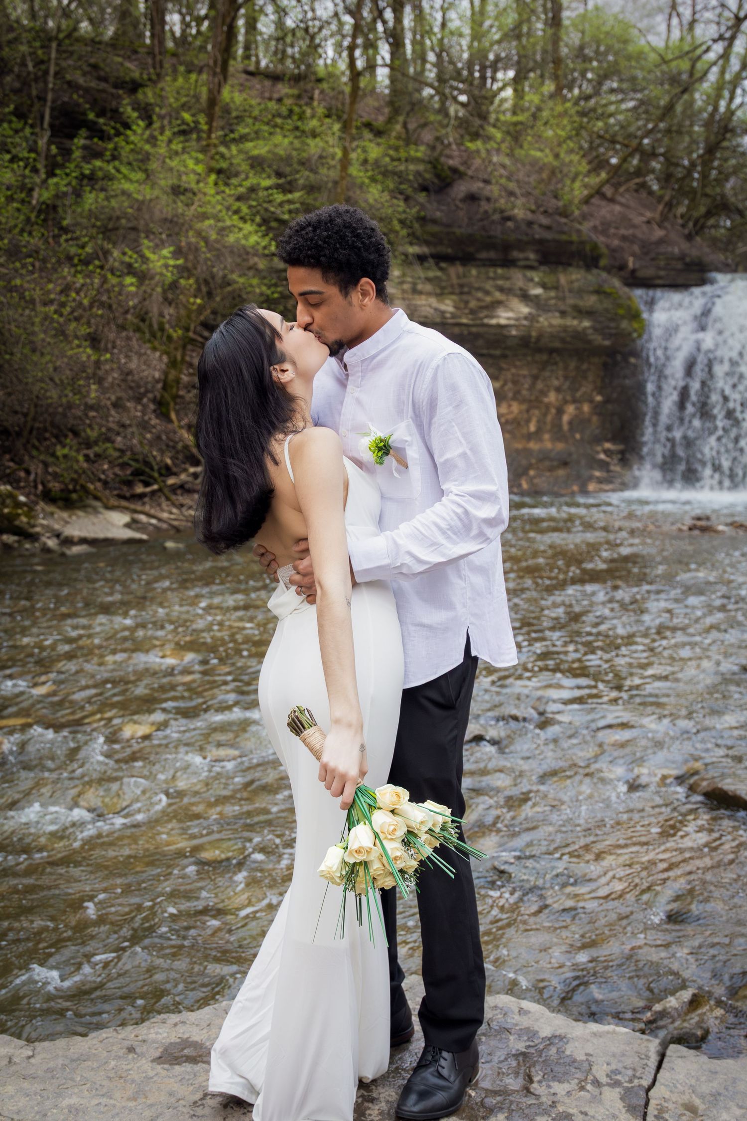 Interracial couple sharing a kiss in wedding attire by a waterfall, bride holding white roses bouquet.