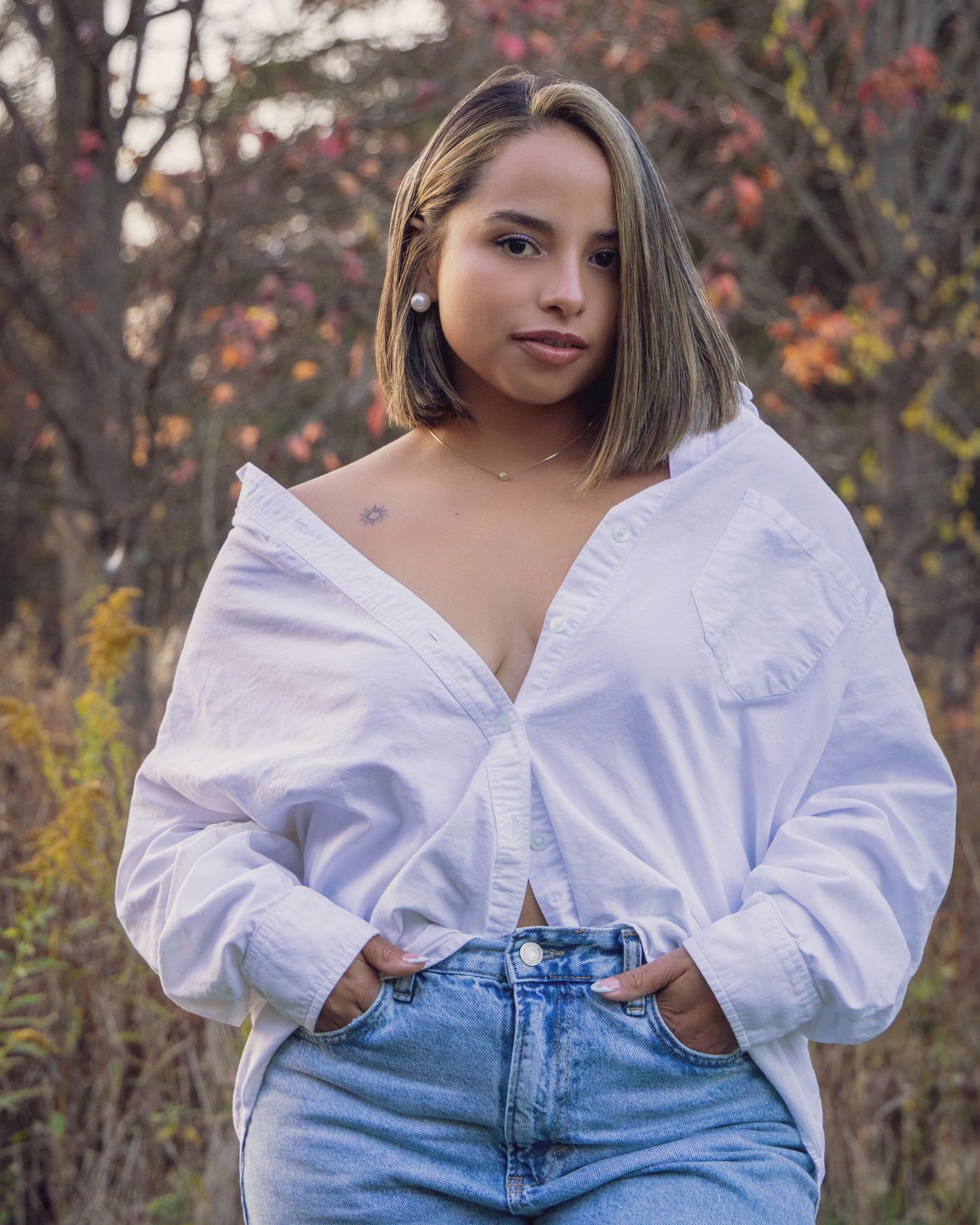 Young woman in white oversized shirt and jeans posing outdoors with autumn foliage background.