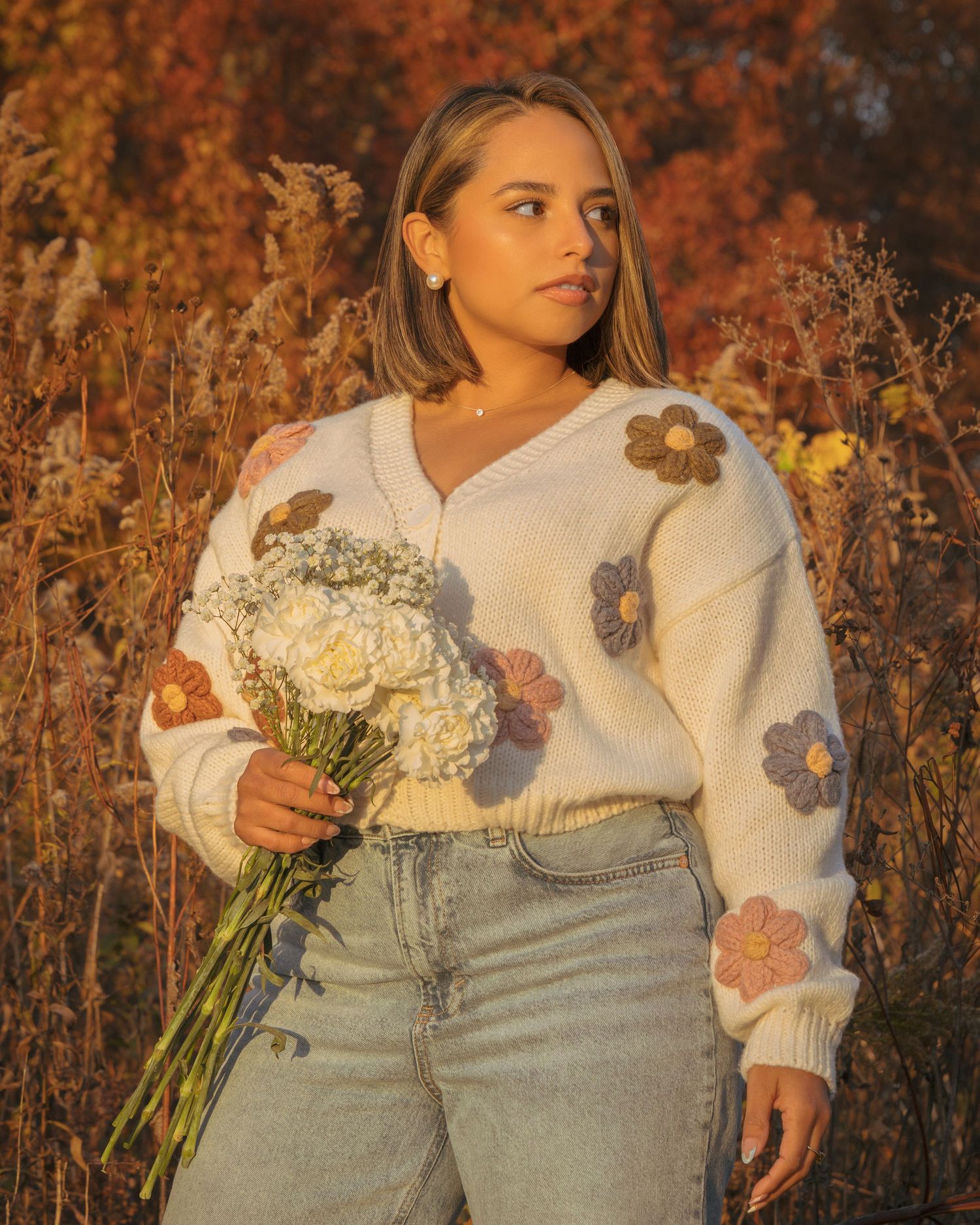 Woman in floral sweater holding white bouquet stands in golden autumn field wearing light jeans.