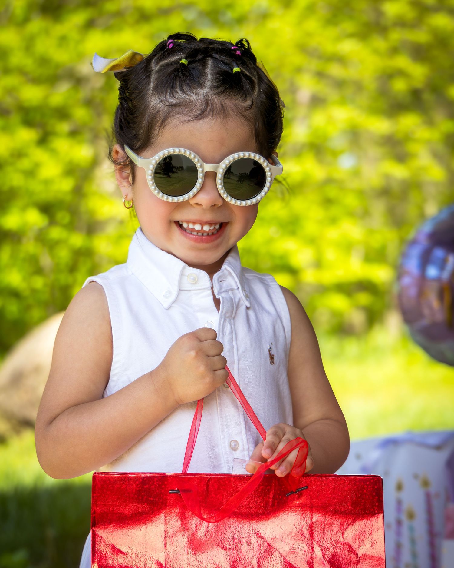 Cheerful child wearing decorative sunglasses and holding red shopping bag in bright outdoor setting.