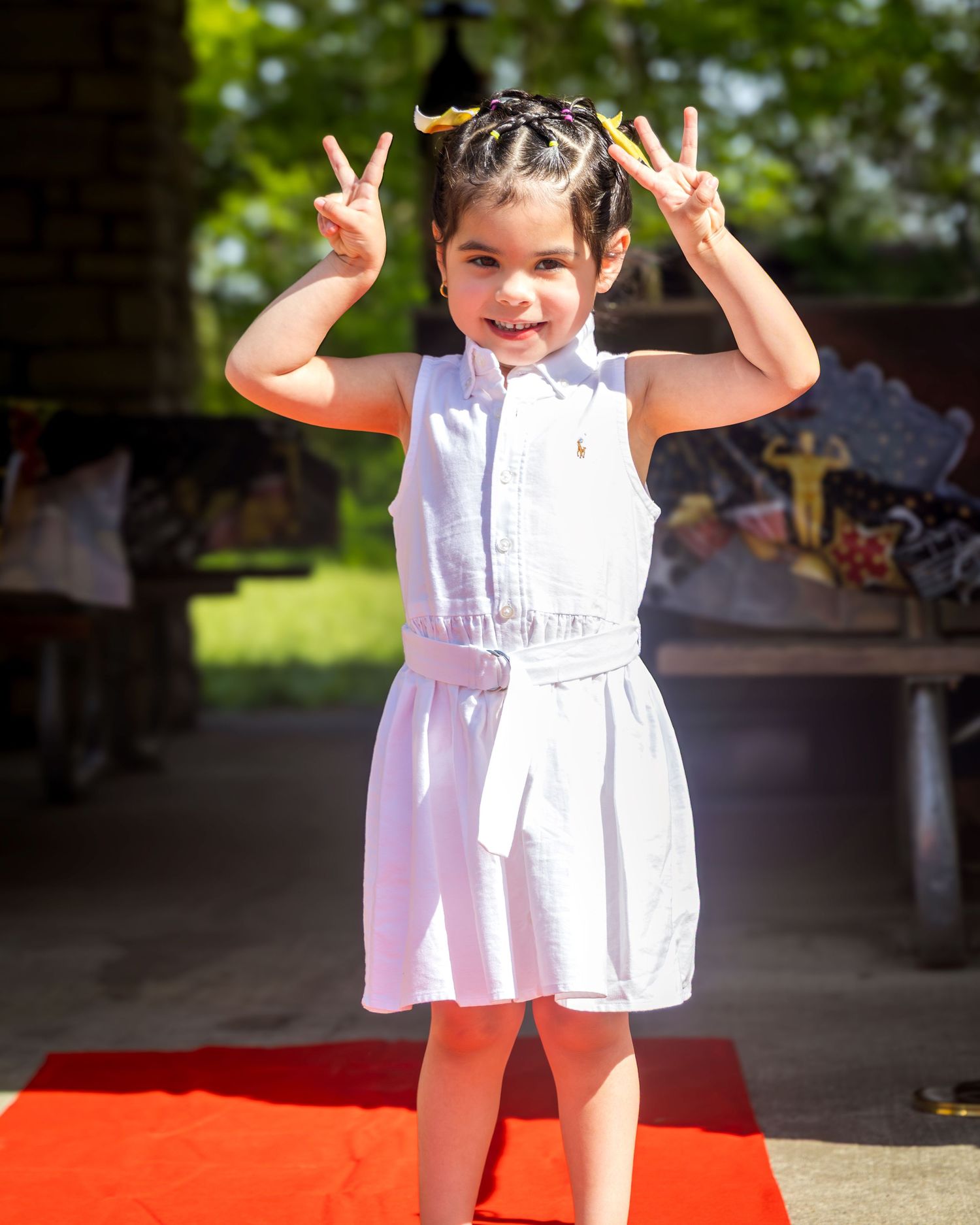 Young girl in white dress making peace signs on red carpet, smiling outdoors in sunny setting.