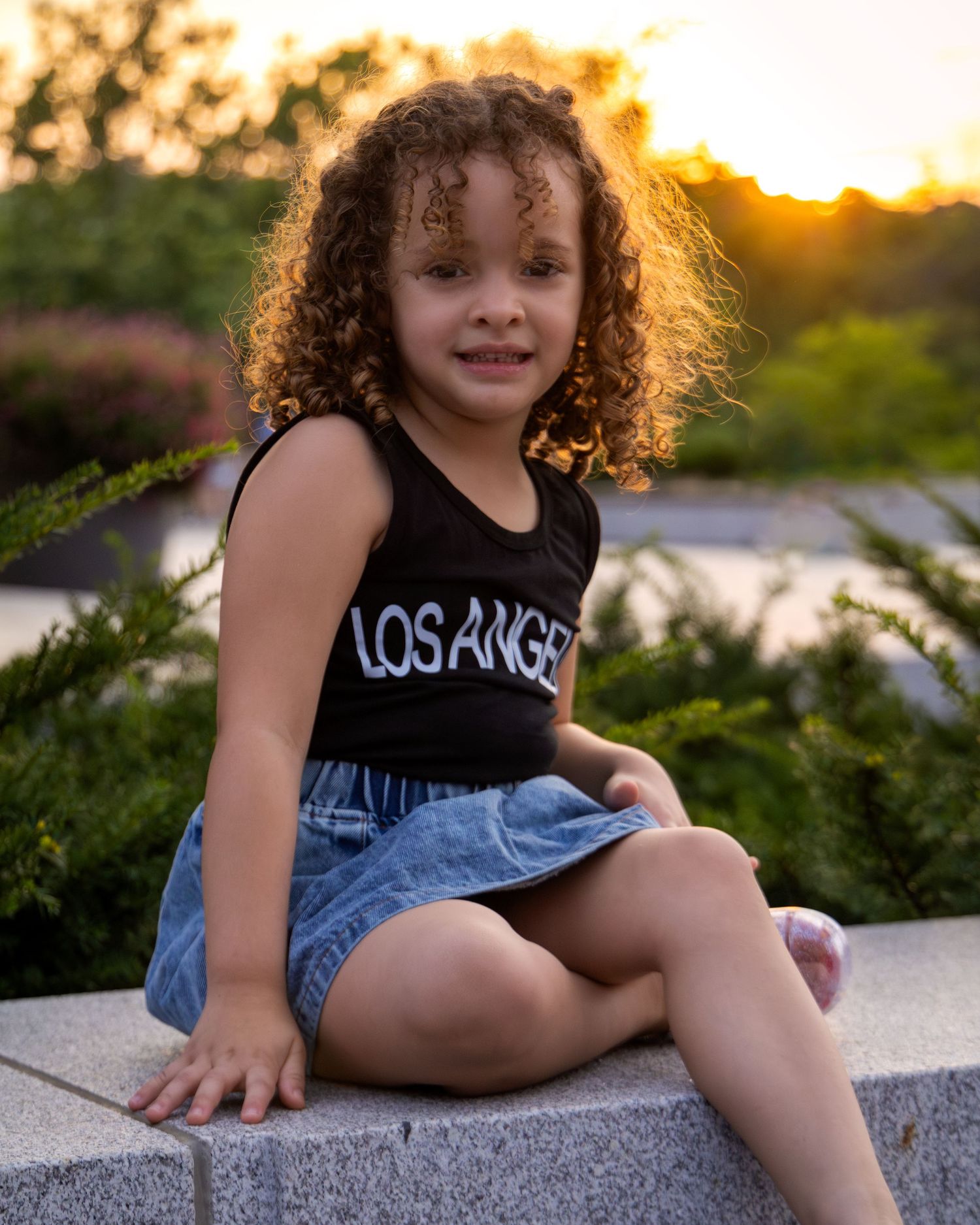 Young girl with curly hair wearing a black Los Angeles tank top and denim skirt sitting outdoors at sunset.