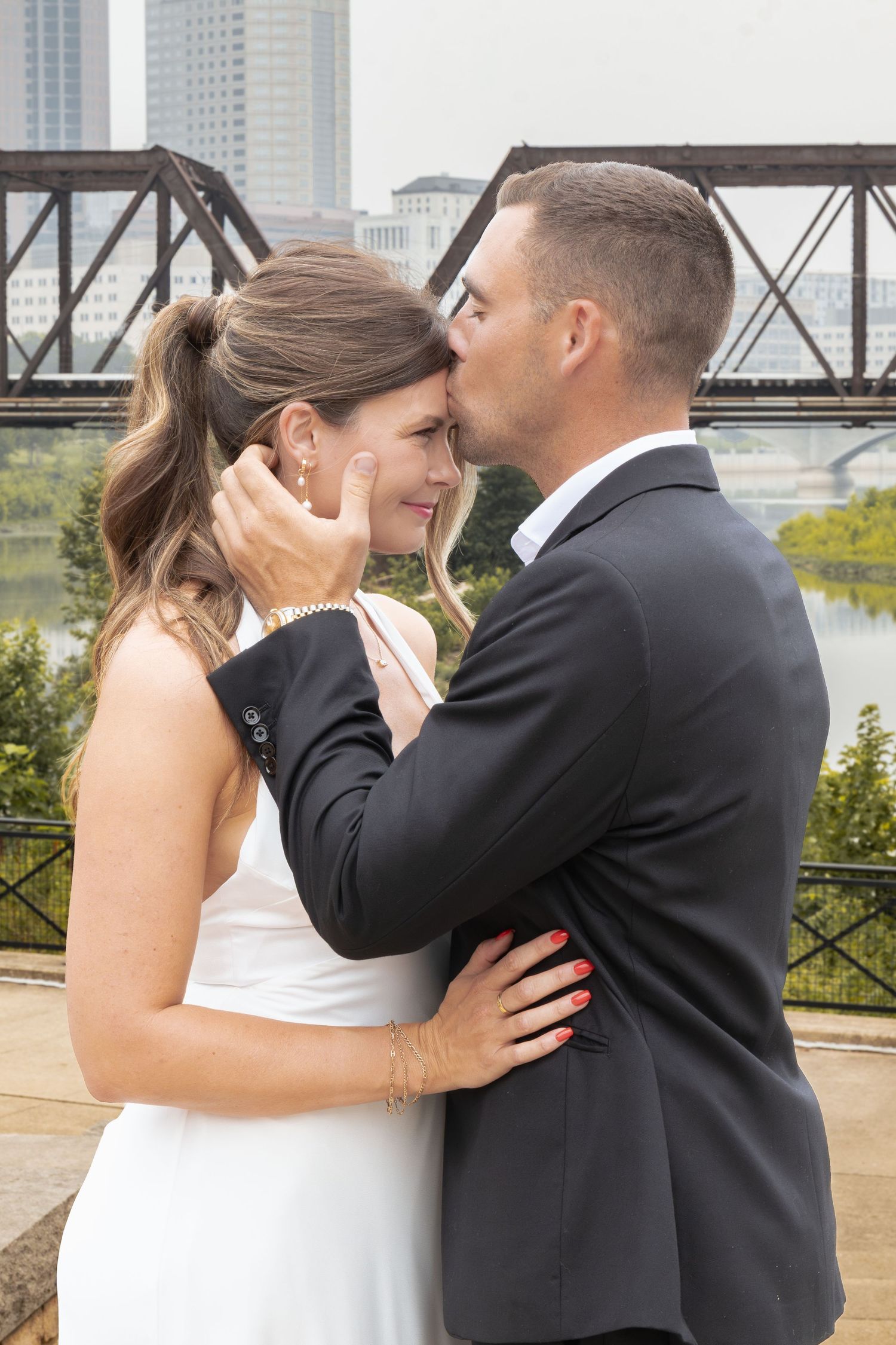 Groom tenderly kisses bride on forehead while holding her face, with a river bridge and city skyline in the background.
