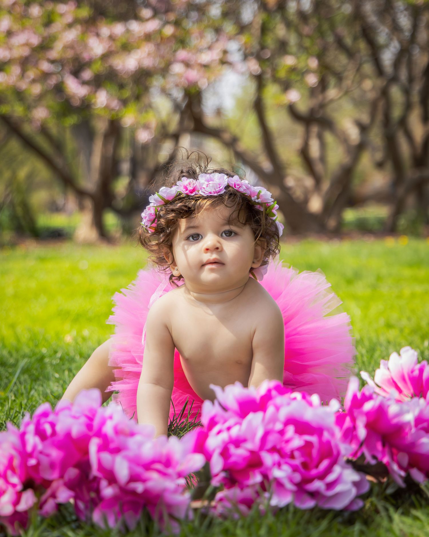 A baby wearing a pink tutu and floral headband poses near vibrant pink flowers in a spring garden setting.
