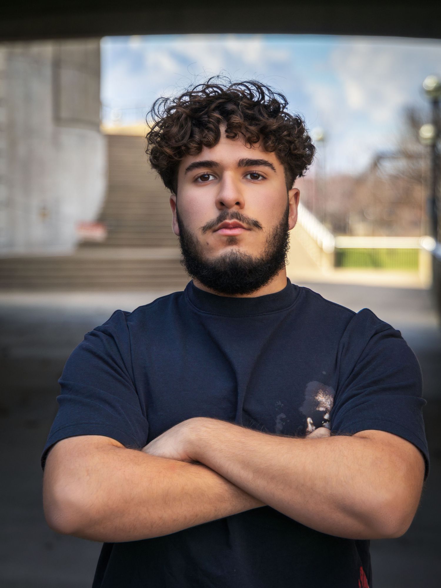 Young man with curly hair and beard stands with arms crossed wearing a dark t-shirt outdoors.