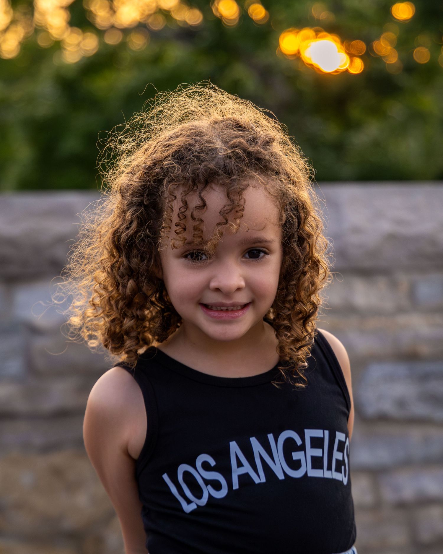 Young girl with curly brown hair wearing a black Los Angeles tank top, smiling outdoors at golden hour.