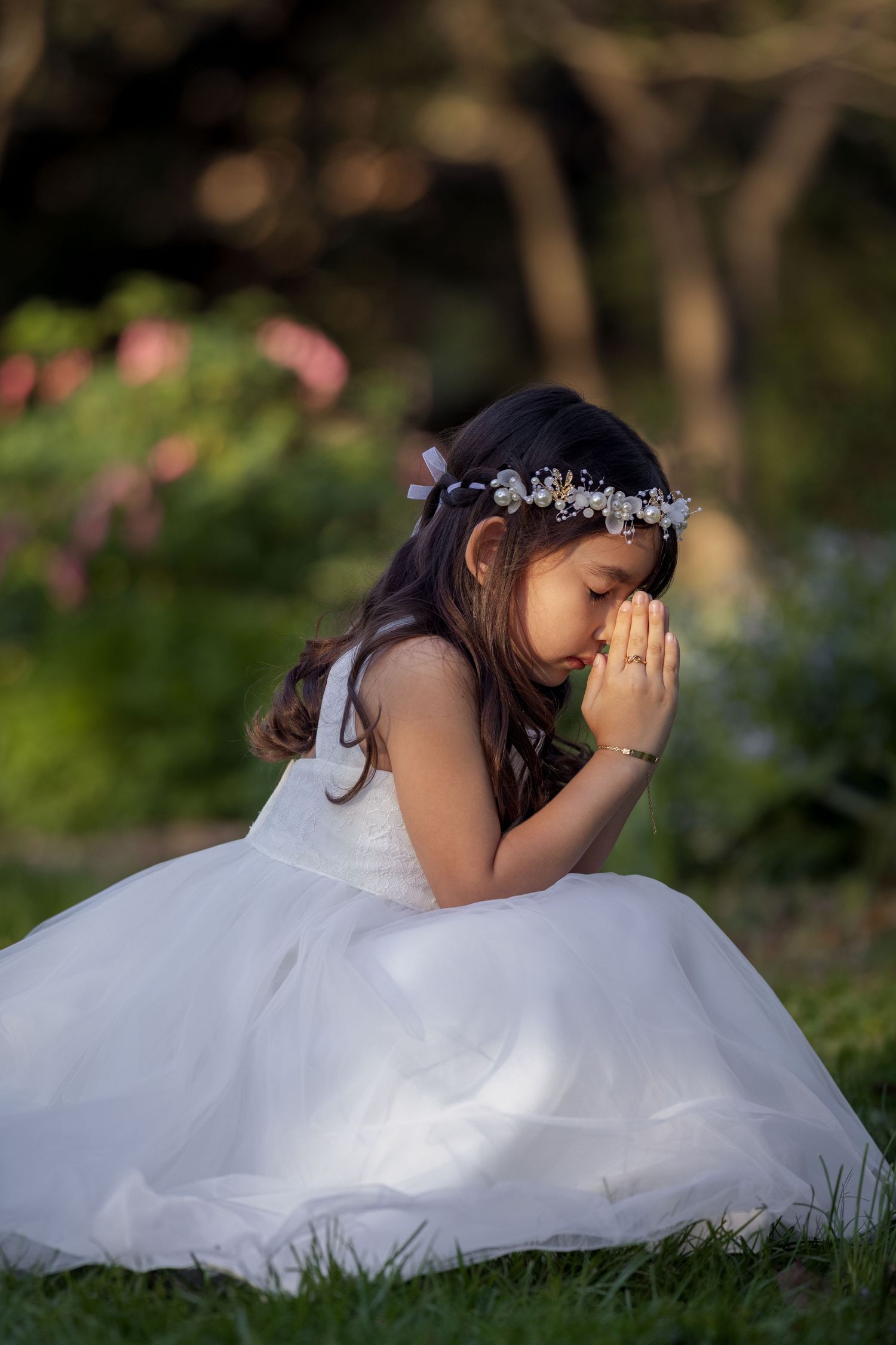 Young girl in white dress and floral crown kneels on grass with hands pressed together in prayer.