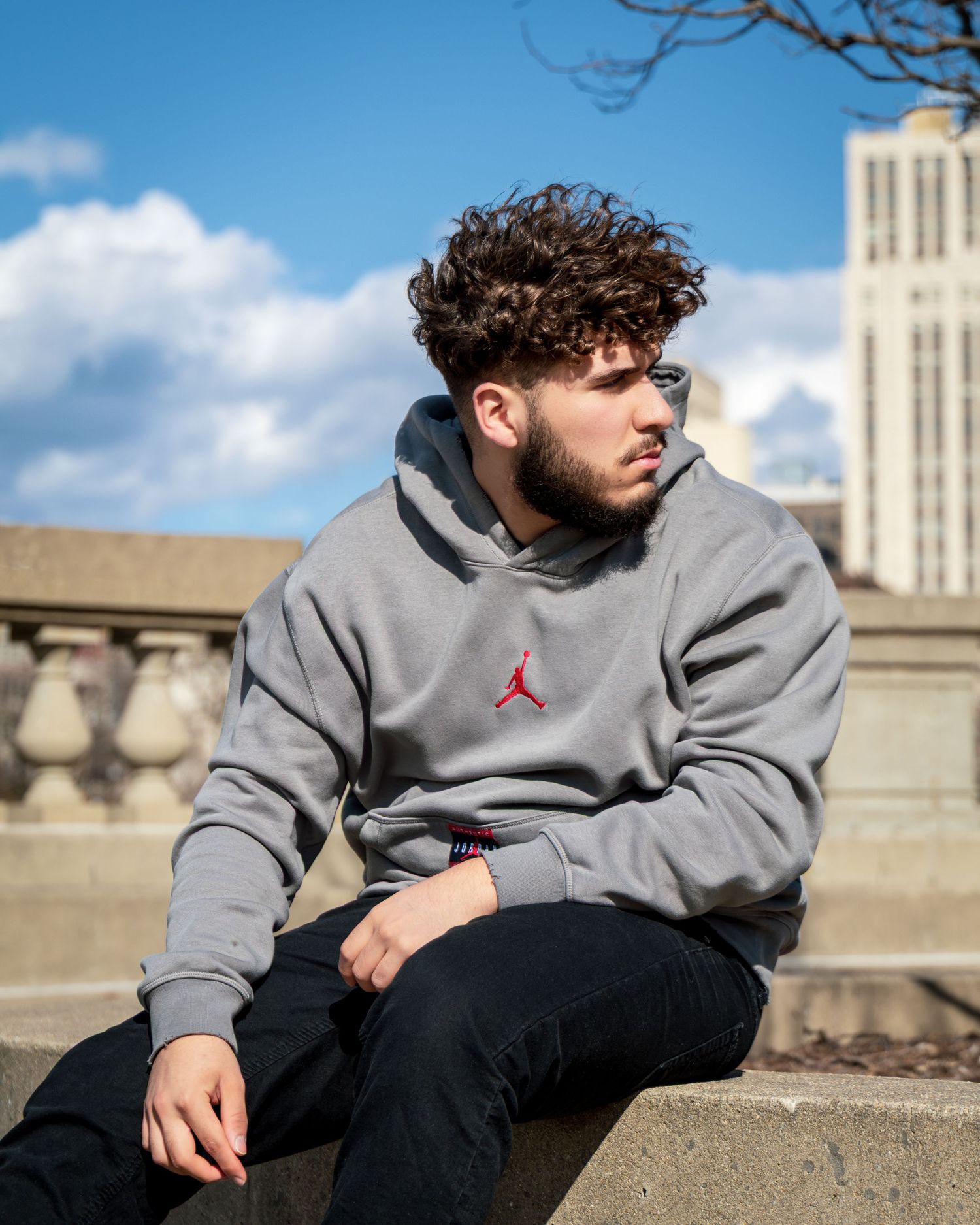 Person in a gray Jordan hoodie sits on stone steps against a blue sky with buildings in background.