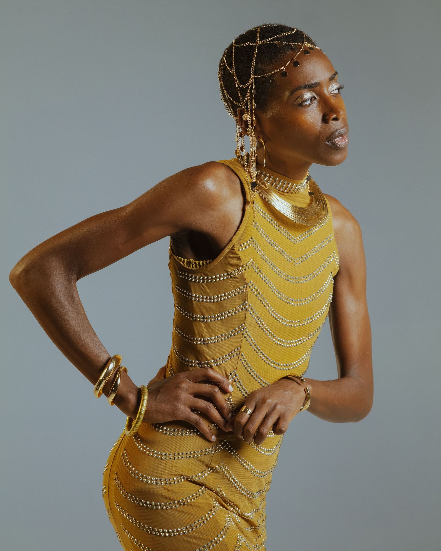 Woman in a gold beaded sleeveless dress with layered necklaces, bracelets and a beaded headpiece posing against a gray studio backdrop.
