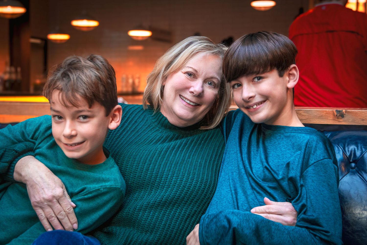 A smiling woman hugs two young boys in green sweaters on a couch.