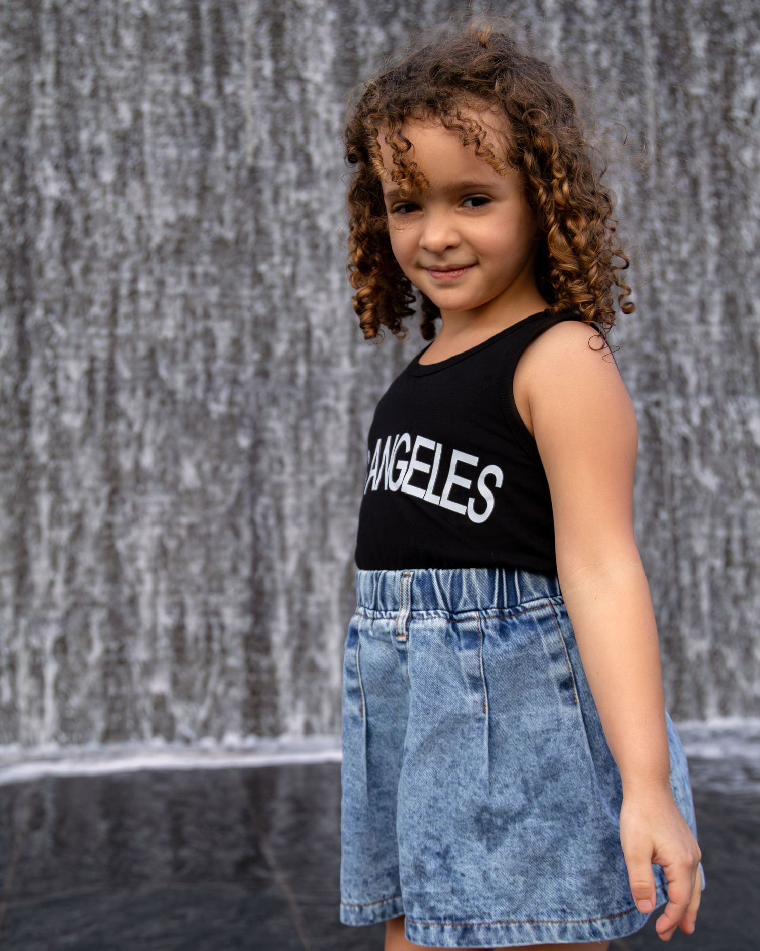 Young girl in black Los Angeles top and denim skirt poses smiling in front of a waterfall backdrop.