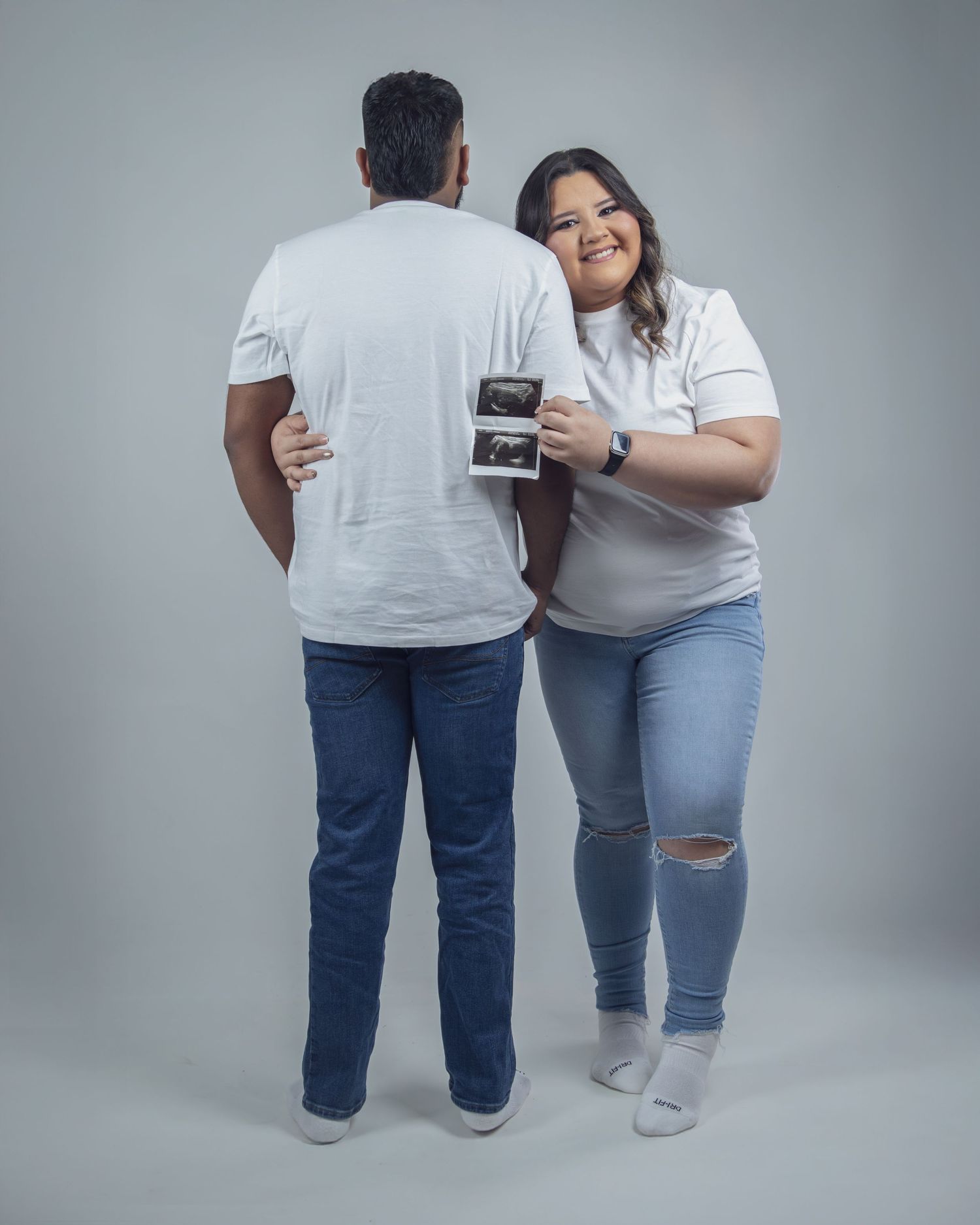 Couple in white tees and jeans holding ultrasound photos, woman smiling at camera during pregnancy announcement shoot.