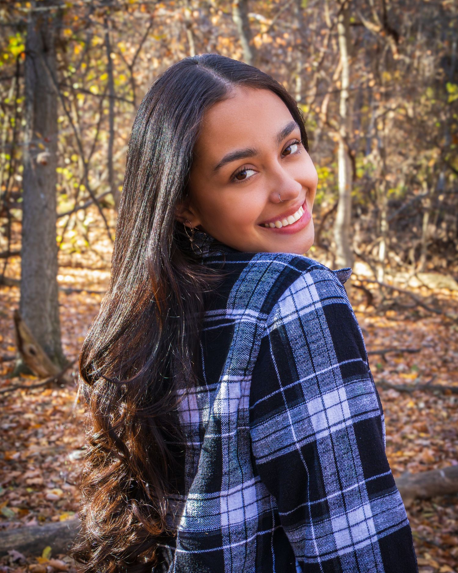 Young woman with long dark hair smiling over shoulder in plaid jacket among autumn forest trees.
