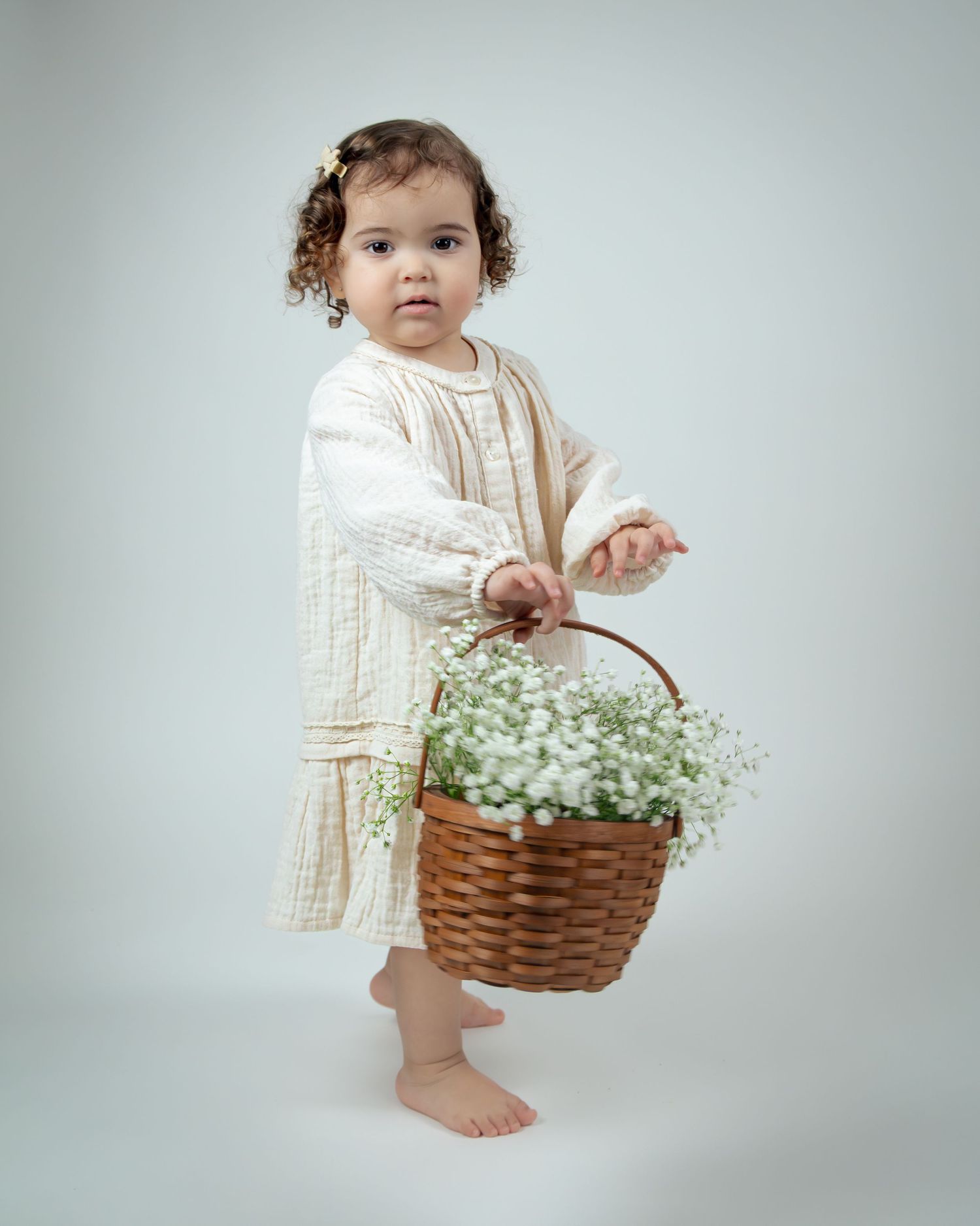 A young child in white lace dress standing next to a woven basket filled with baby's breath flowers.