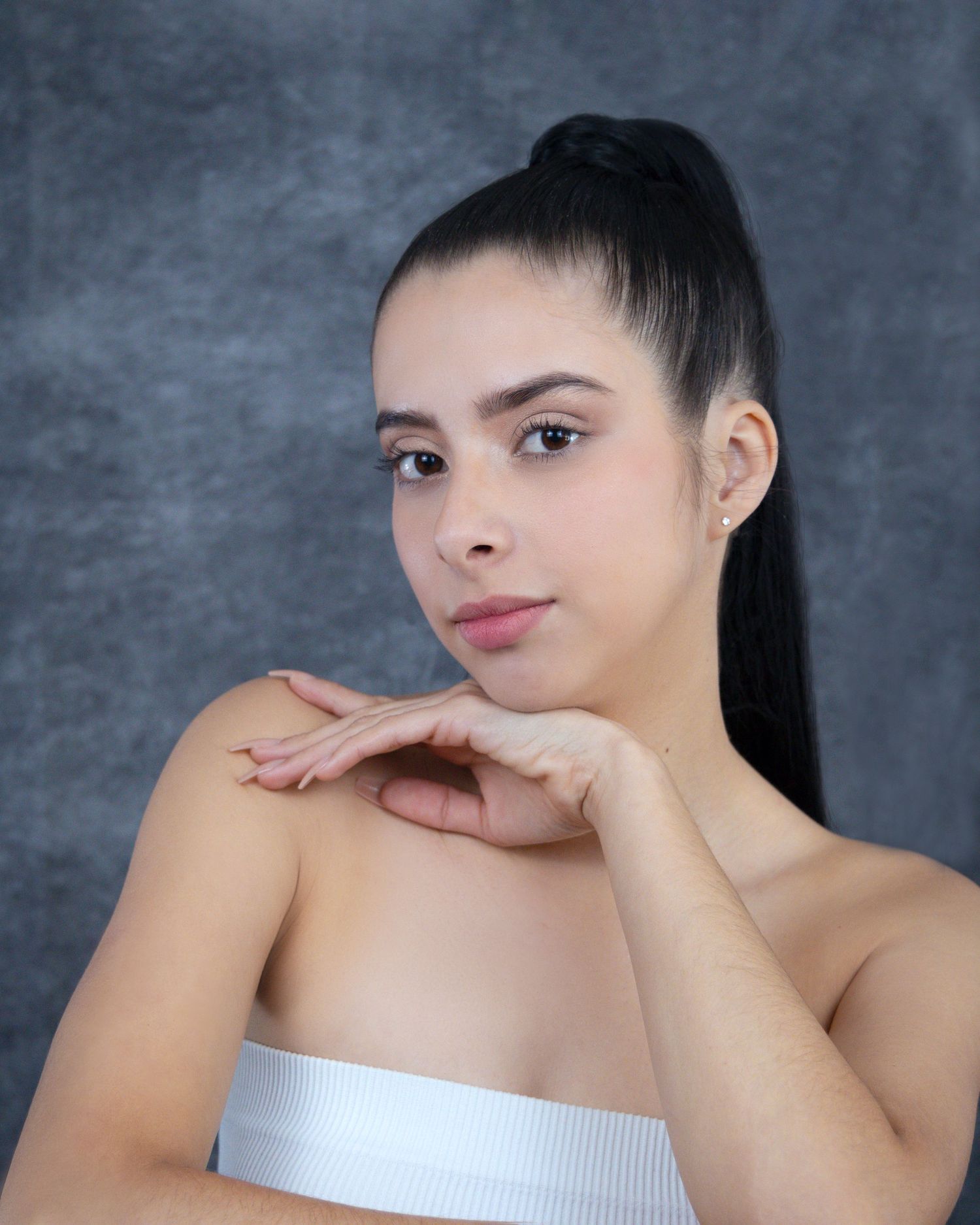 Young woman with high ponytail posing gracefully, resting chin on hands, wearing white tube top against gray backdrop.