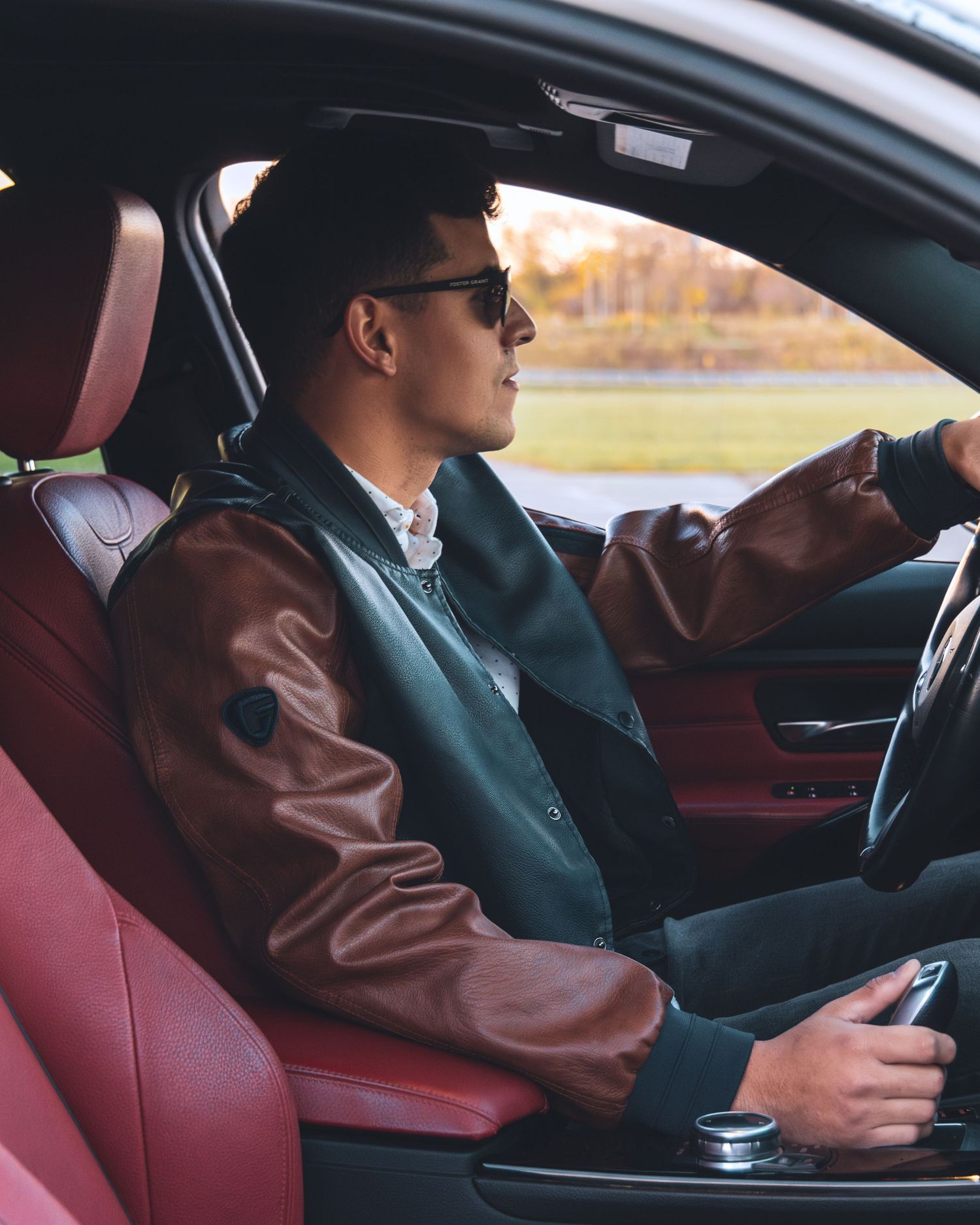 Young man in leather varsity jacket and sunglasses driving a luxury car with red interior.