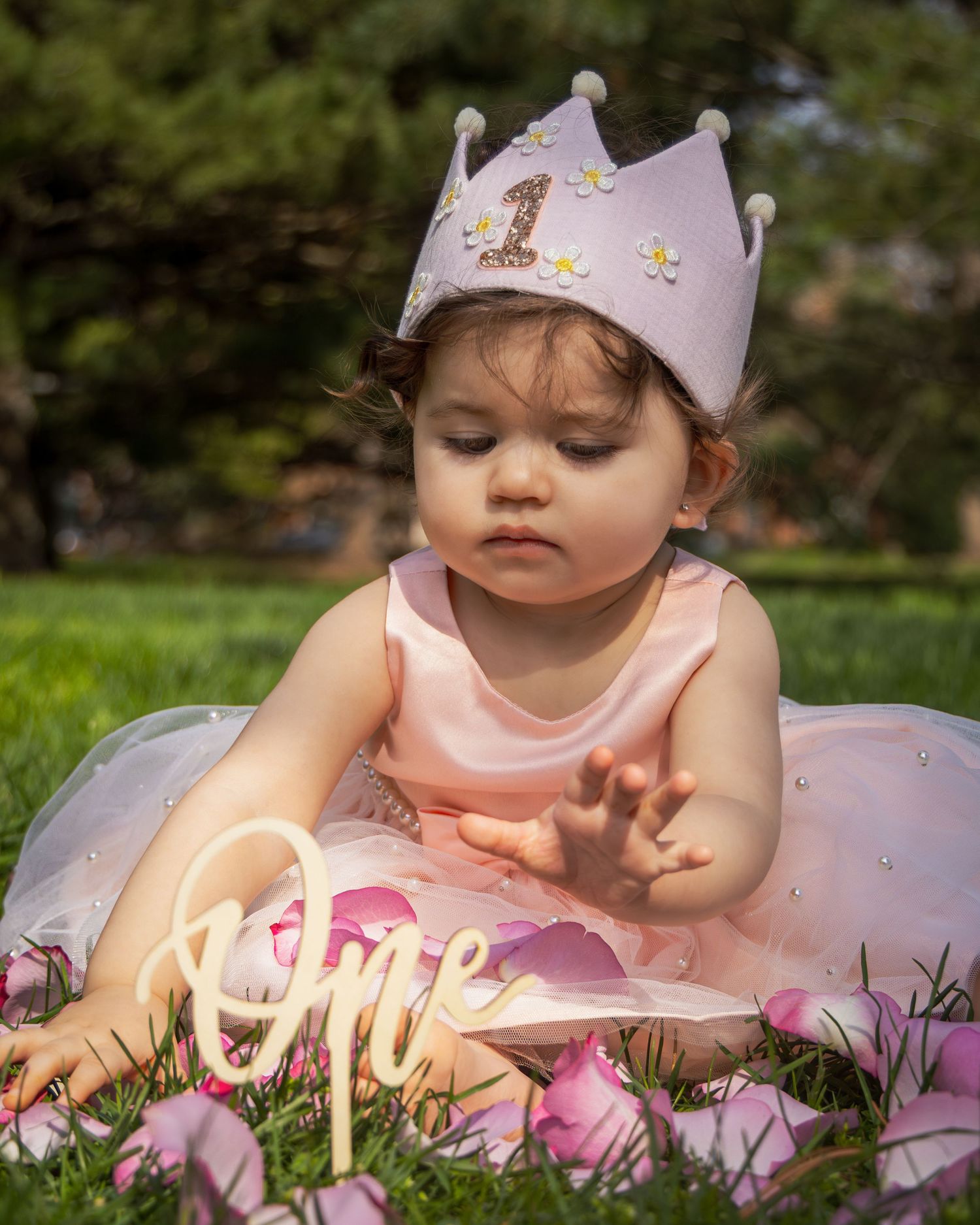 Baby in pink dress and crown sitting outdoors with illuminated 'One' sign and flowers.