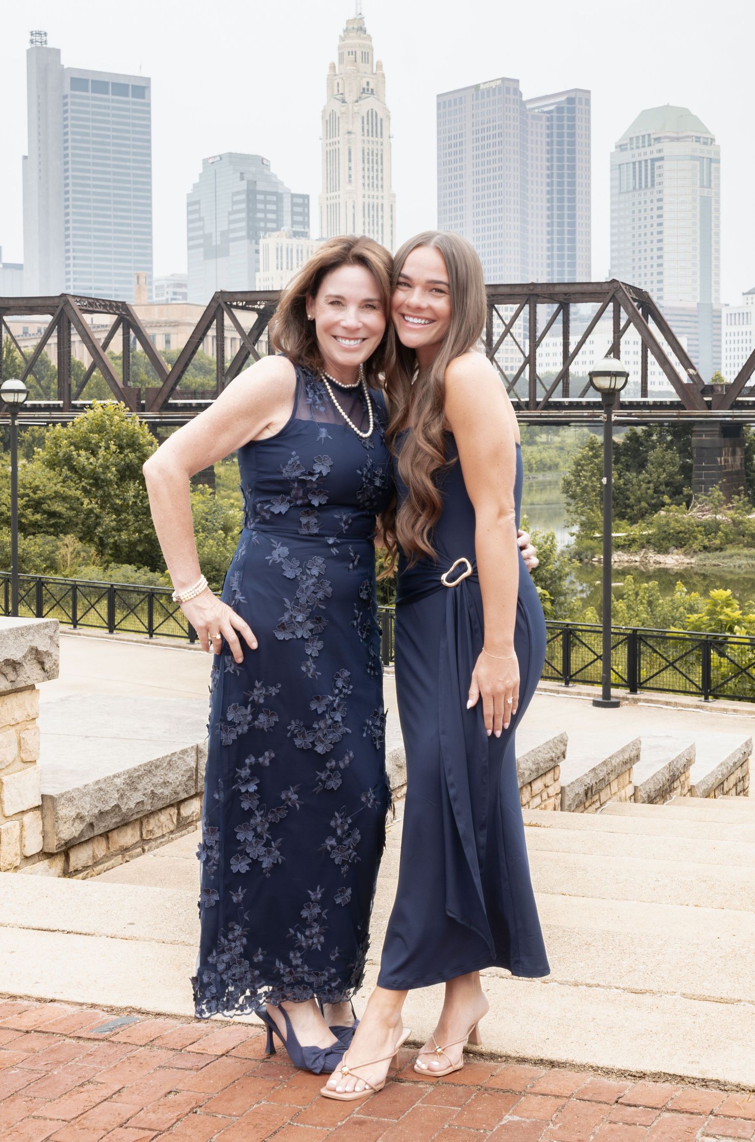 Two women in navy blue dresses pose together with a city skyline and bridge in the background.