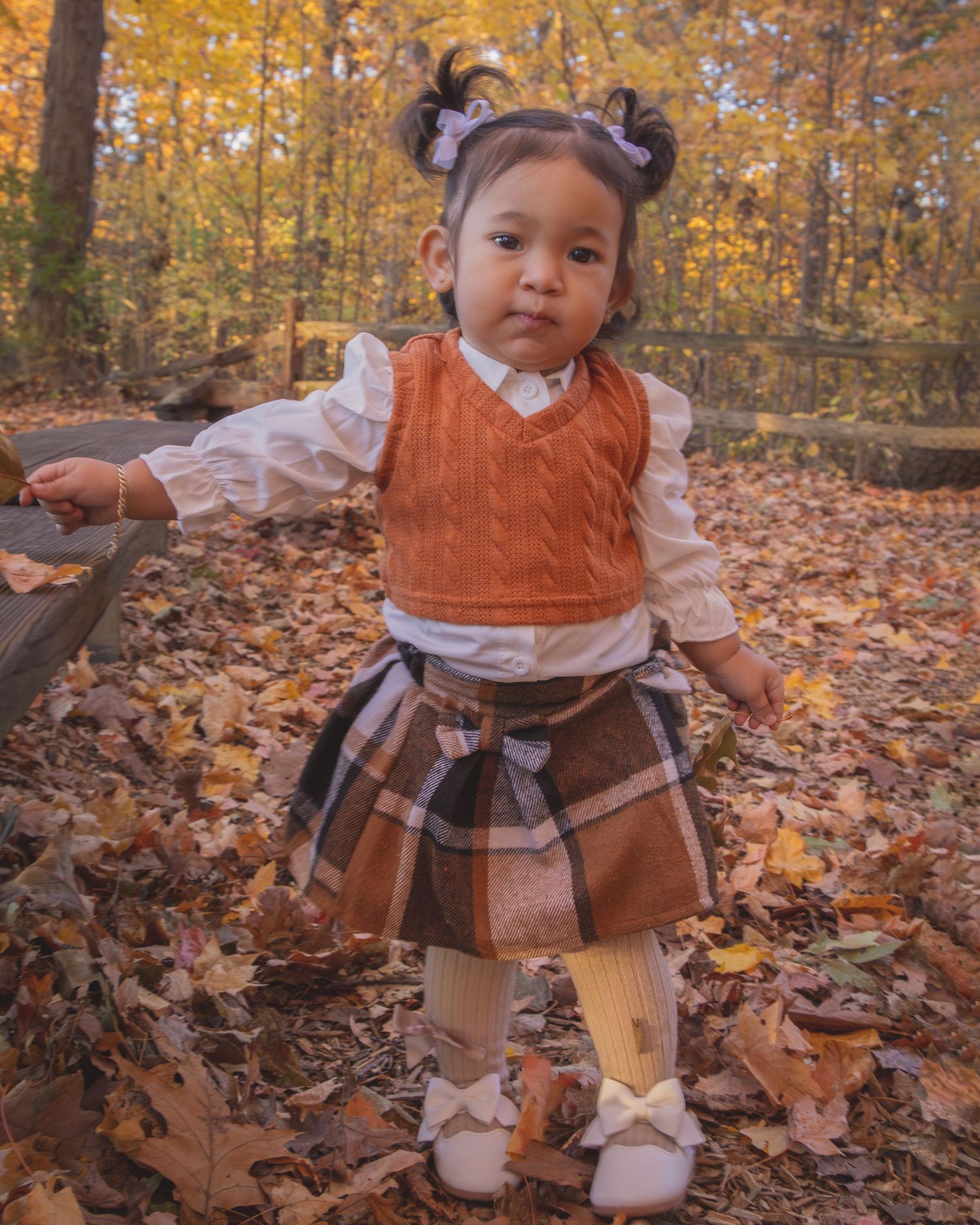 Toddler girl in autumn plaid skirt and orange vest poses among fall leaves in a forest setting.