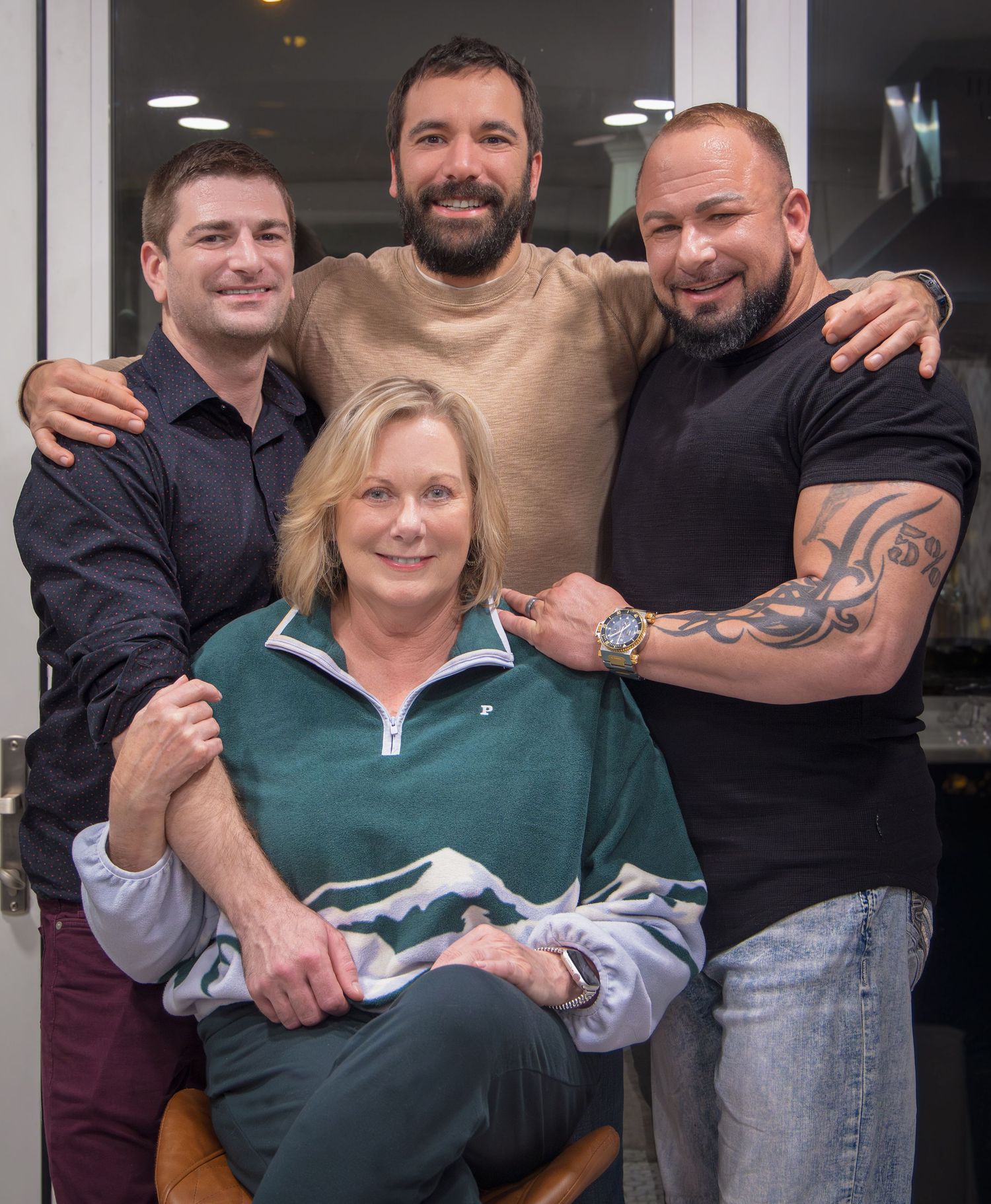 Four people pose together smiling, three men standing behind a seated blonde woman in a green pullover.