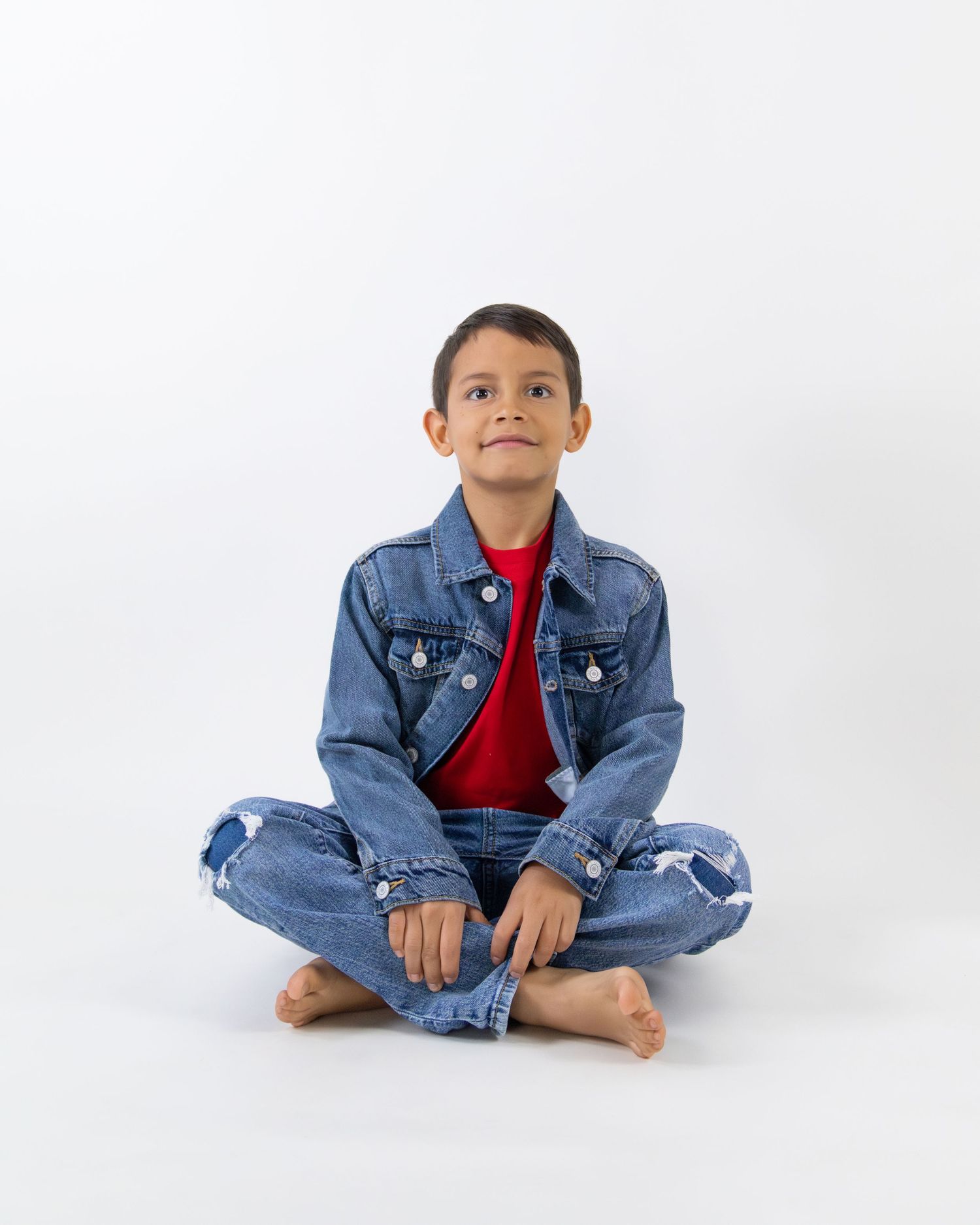 A young boy sits cross-legged barefoot, wearing a denim jacket, red shirt, and ripped jeans, looking up.