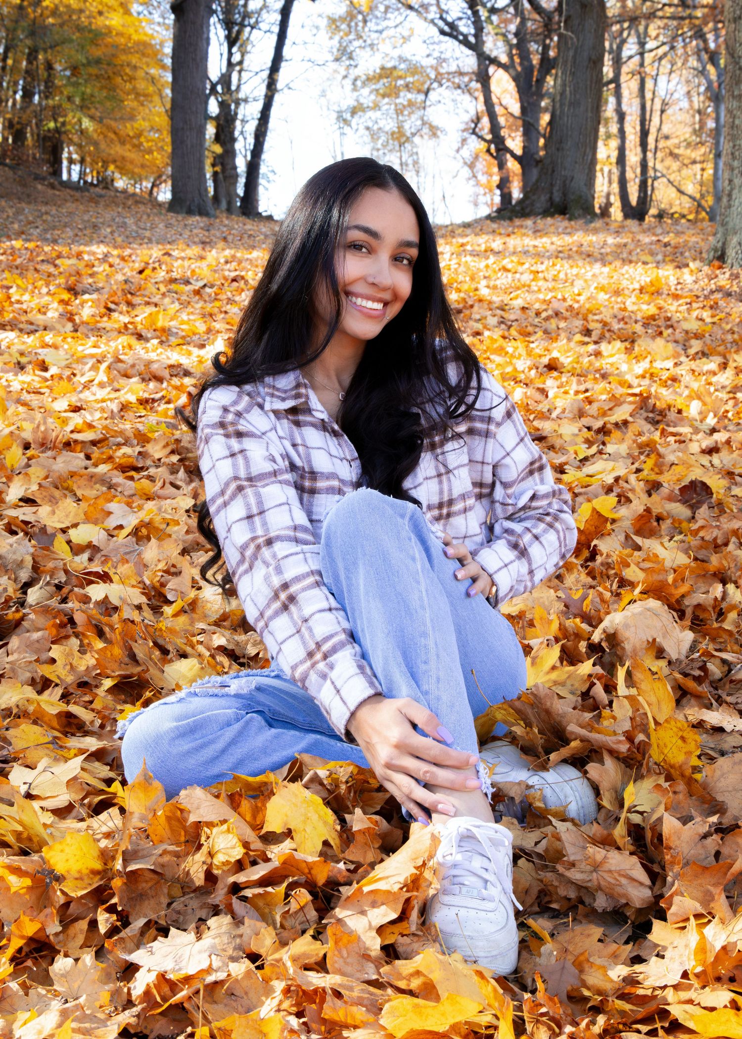 Young woman smiling in autumn park, sitting on fallen leaves wearing plaid shirt and jeans.