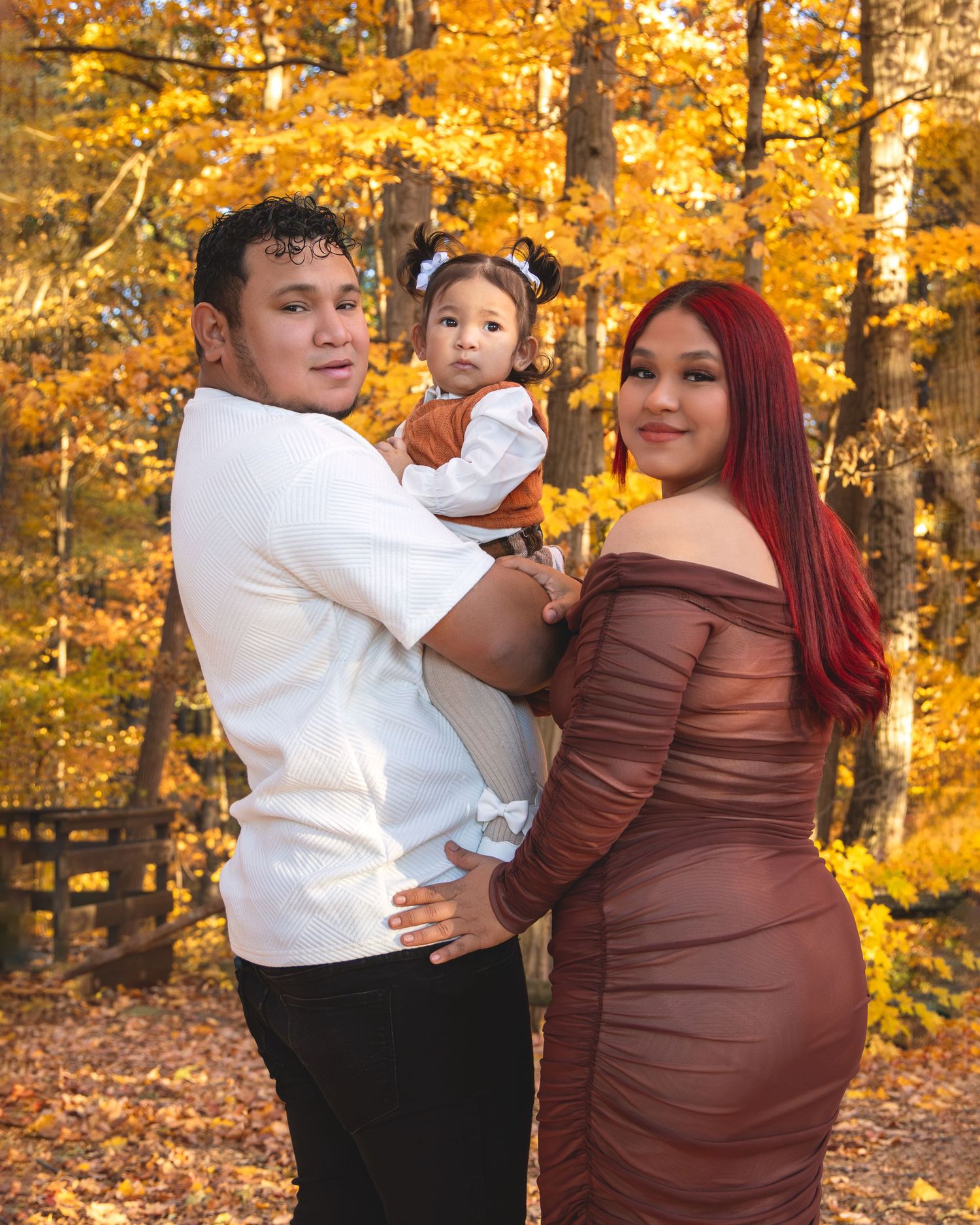 Young family poses together in autumn forest with golden foliage, holding their toddler in fall outfits.
